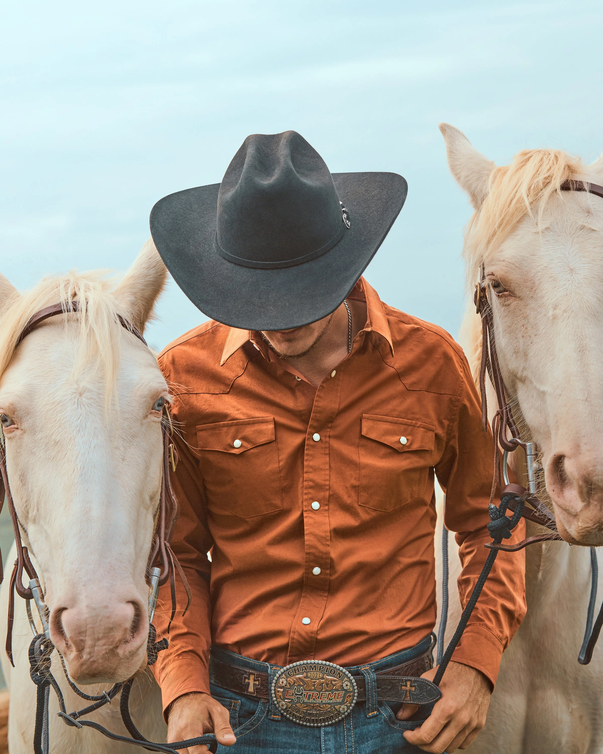 A man wearing a black wide-brimmed hat, an orange shirt, and a large belt buckle stands between two white horses, with his head bowed and not visible.