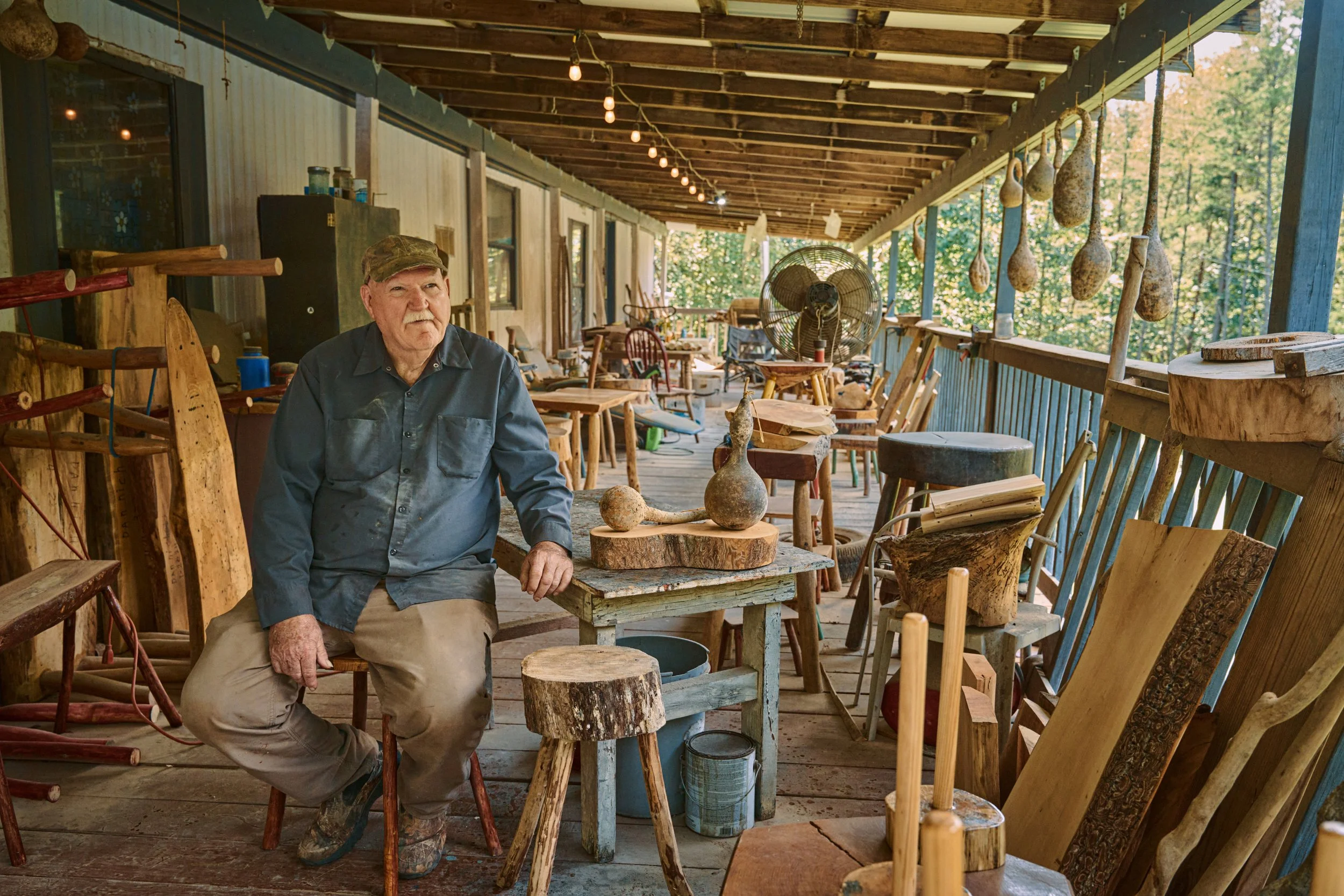 An elderly man sitting on a chair in a rustic woodworking workshop surrounded by handmade wooden objects and tools, with a porch view of trees in the background.