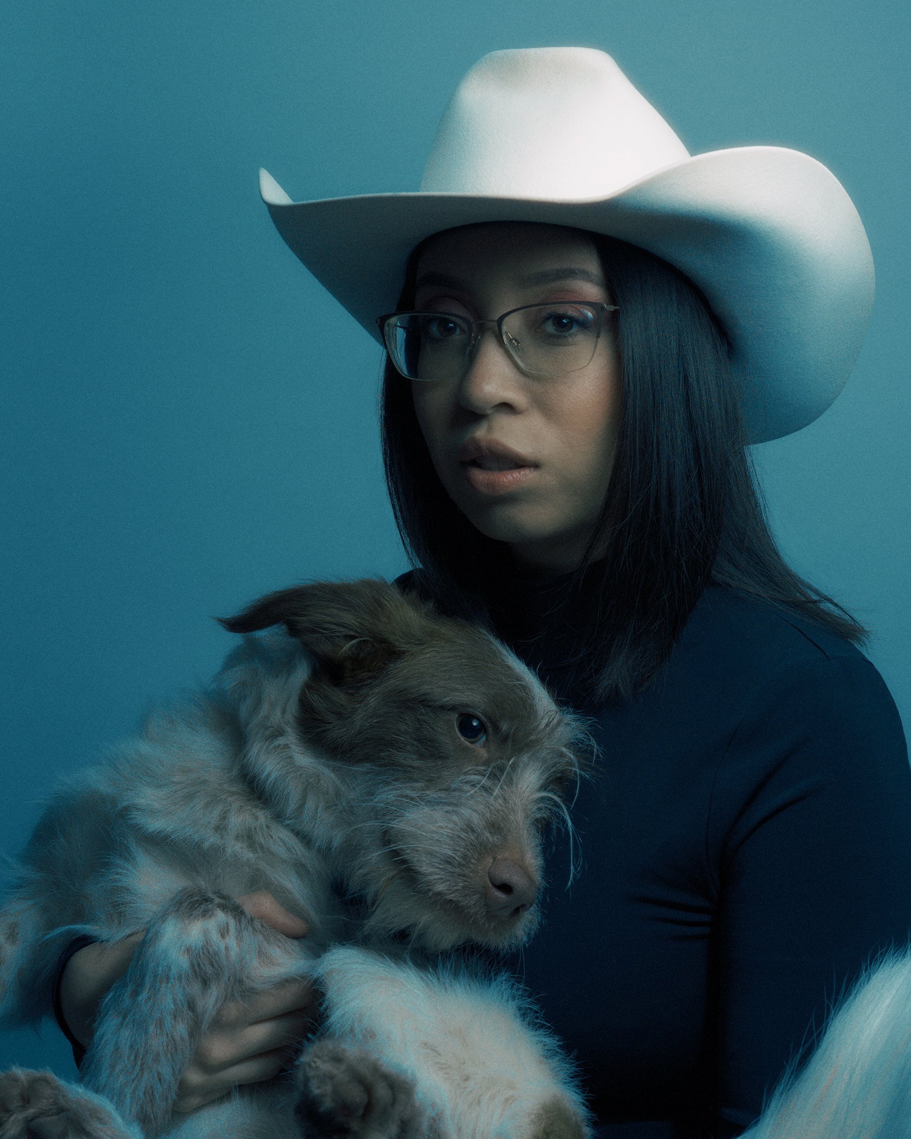 A woman wearing glasses and a white cowboy hat holds a brown and white dog against a light blue background.