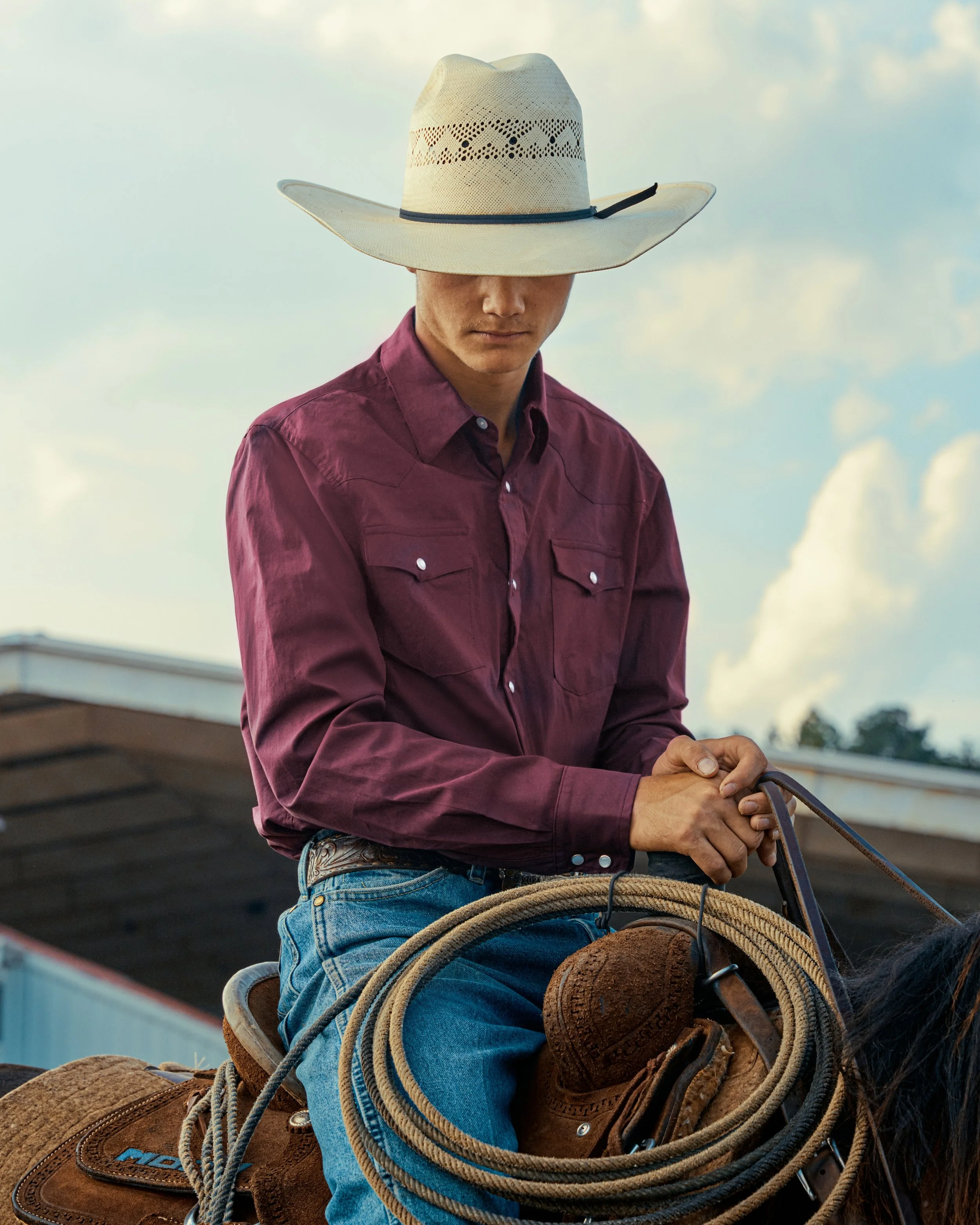 A man wearing a wide-brimmed hat, a maroon long-sleeve button-up shirt, and jeans, sitting on a horse with a lasso, under a partly cloudy sky.
