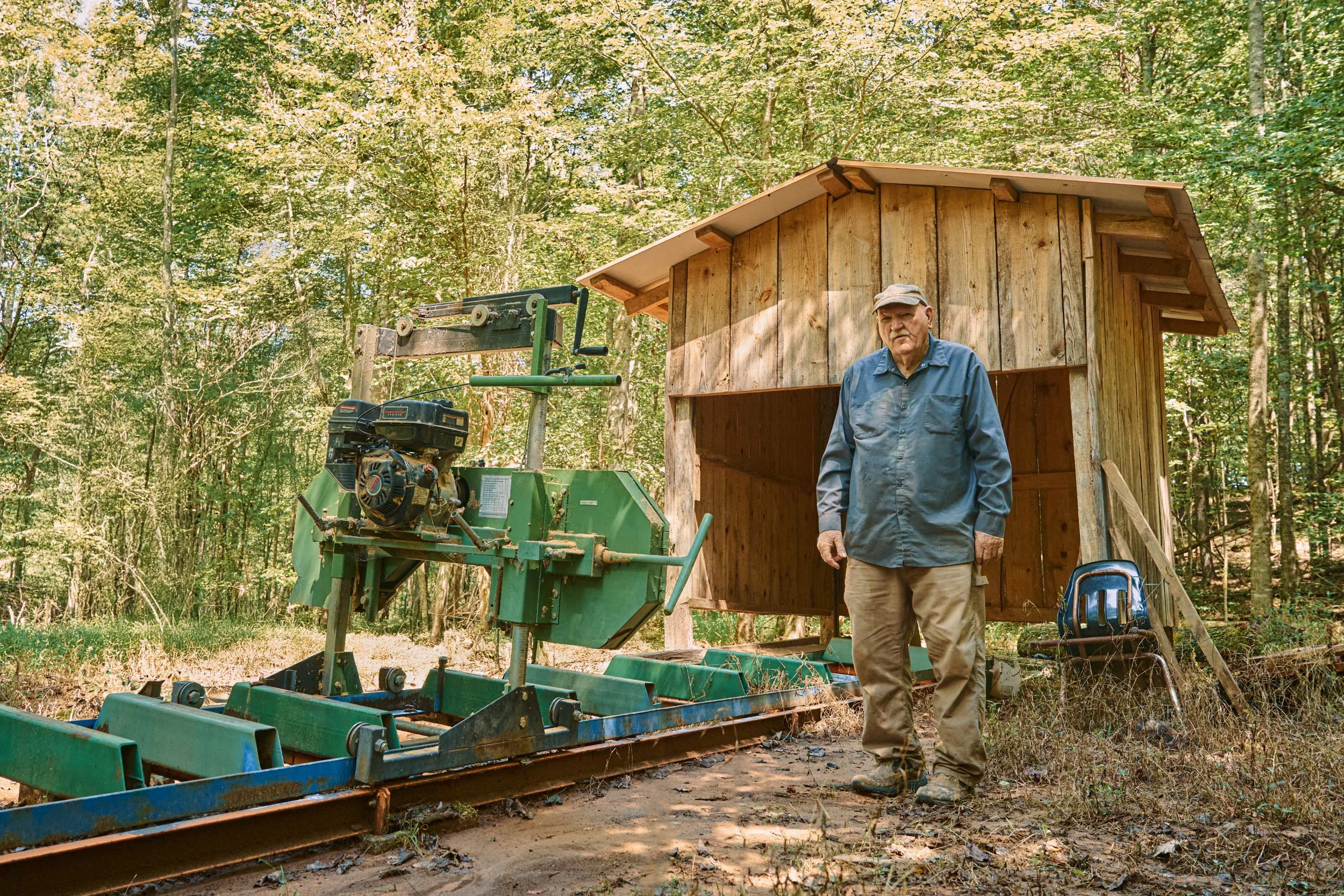 An elderly man standing in front of a wooden shed in a wooded area, with a rototiller or similar gardening equipment nearby.