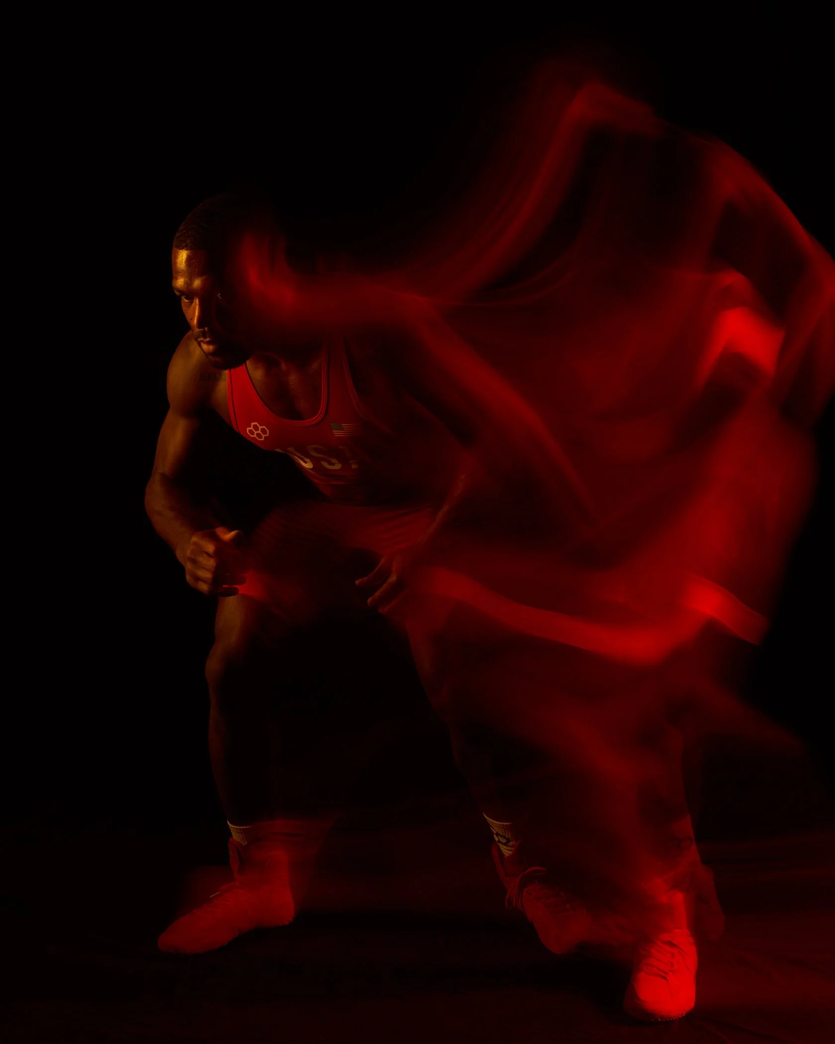 An athlete in a red uniform crouches in a starting position with a dark background, illuminated with red lighting effects creating motion blur.