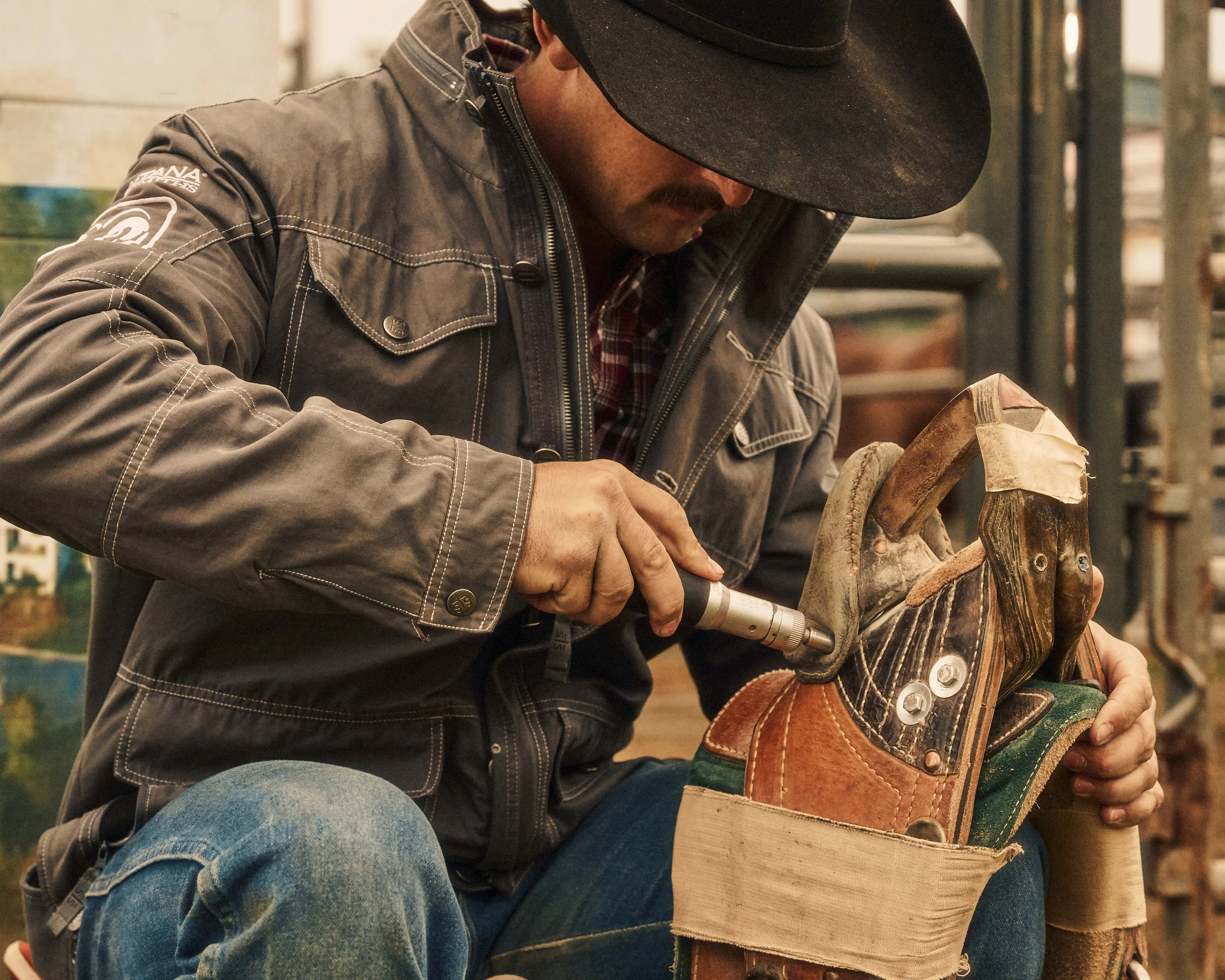 A man wearing a black hat and gray jacket is repairing a cowboy boot with a handheld tool in a workshop.