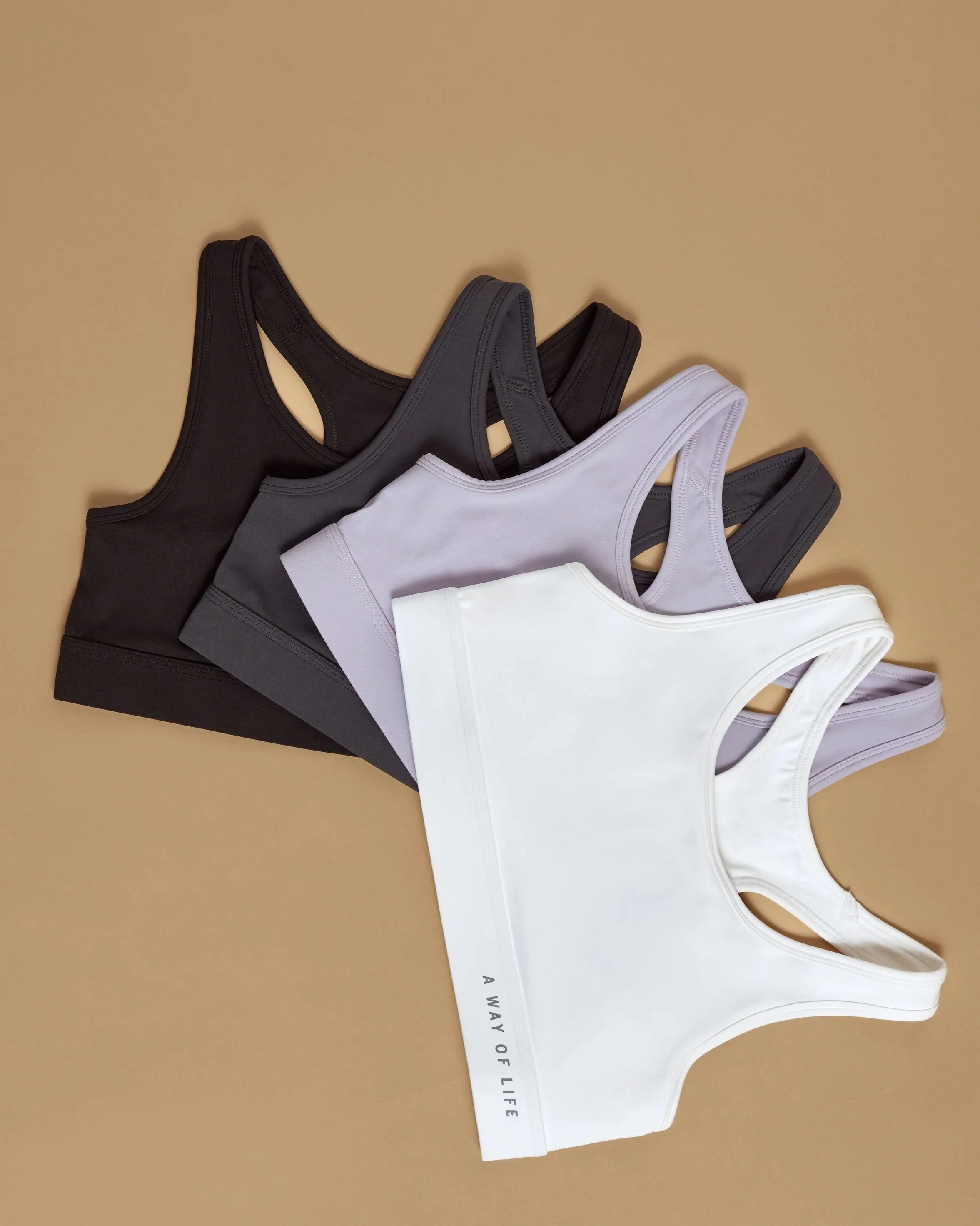 A collection of four sports bras in black, gray, lavender, and white, arranged on a beige surface.