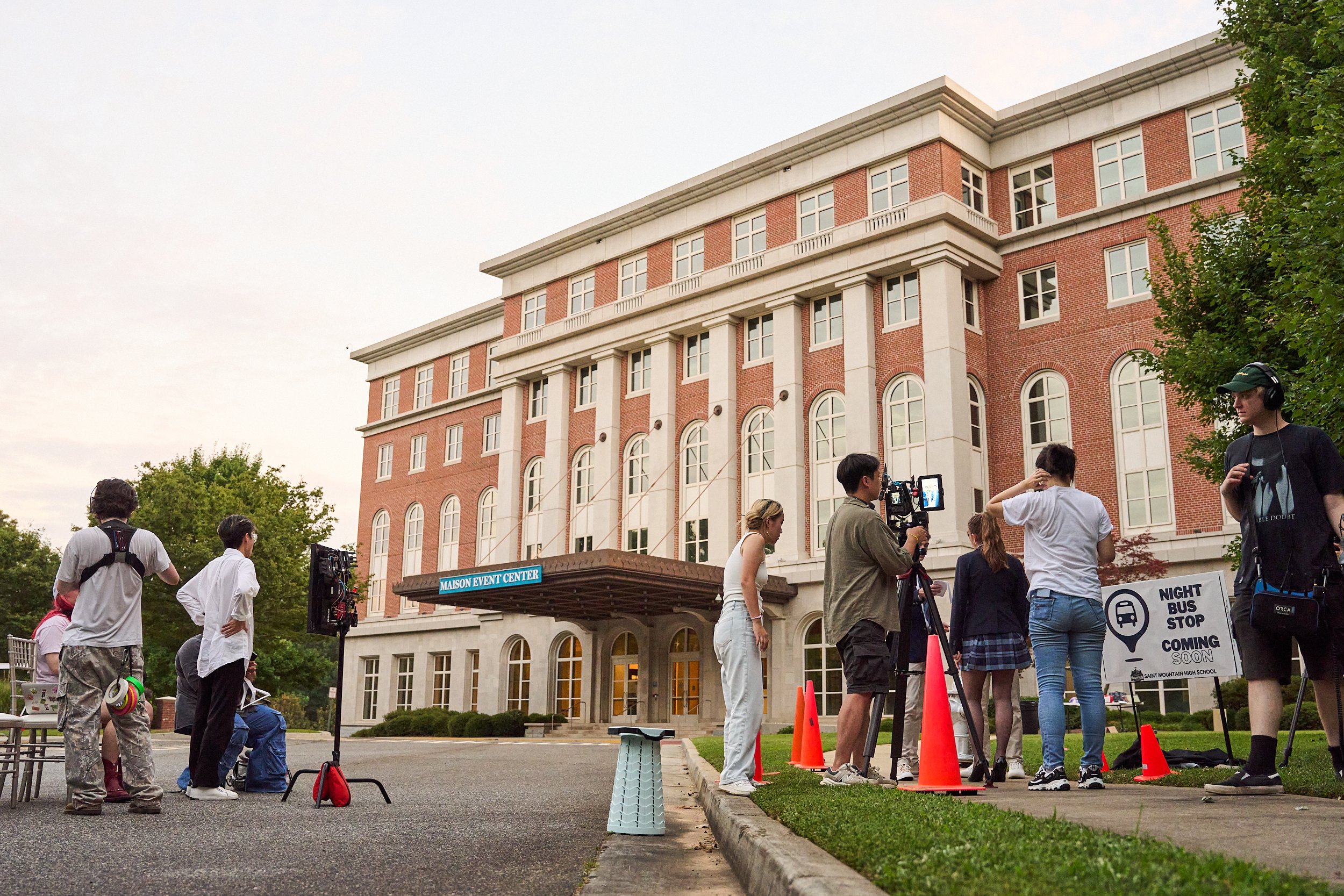 Filming crew setting up outside the Maison Event Center at night, with people preparing for a shoot, a sign indicating a night bus stop coming soon, and lighting equipment on a city street.