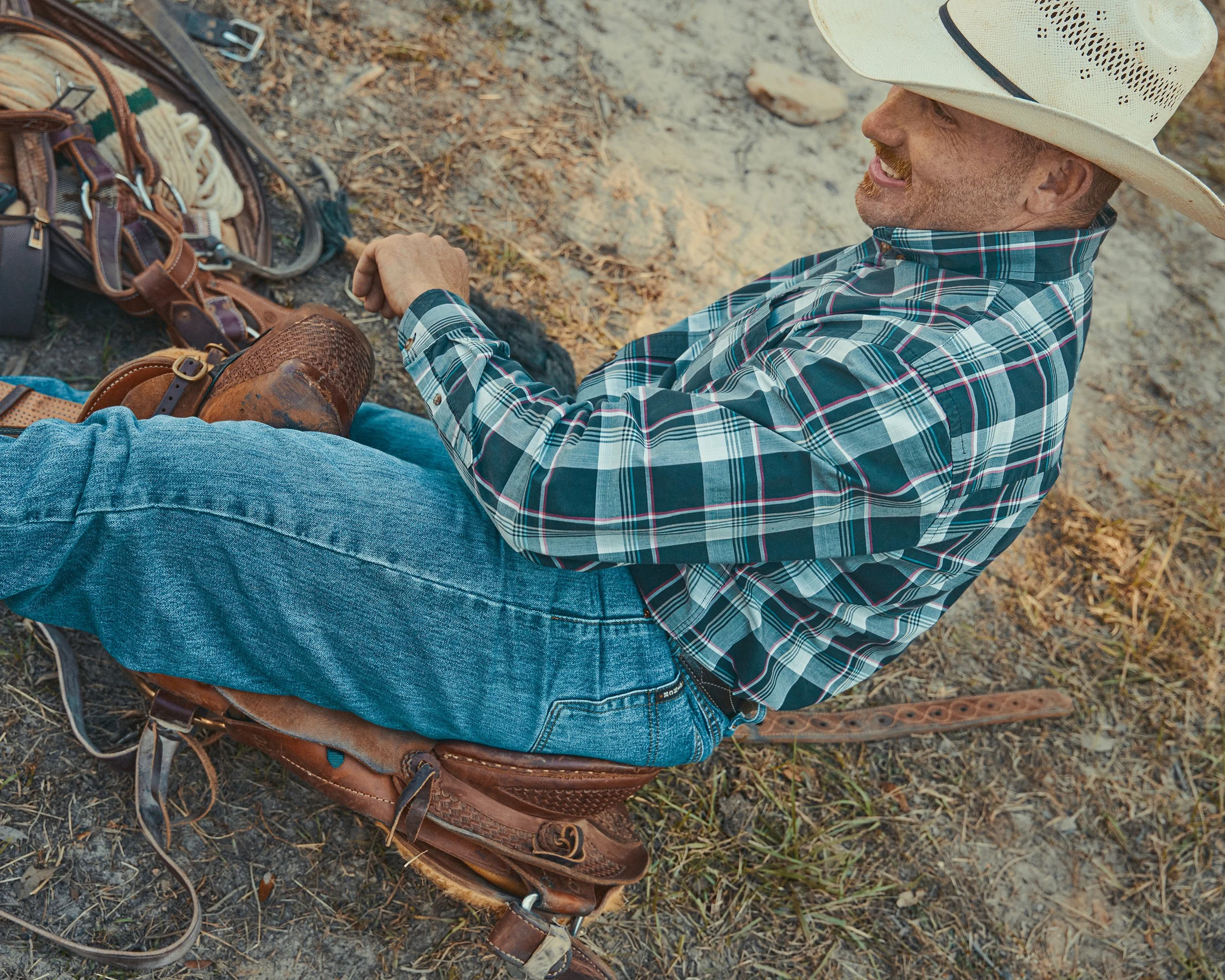 A man sitting on a saddle, smiling, wearing a plaid shirt, blue jeans, cowboy hat, and cowboy boots. There are leather gear and rope nearby, on the ground outdoors.
