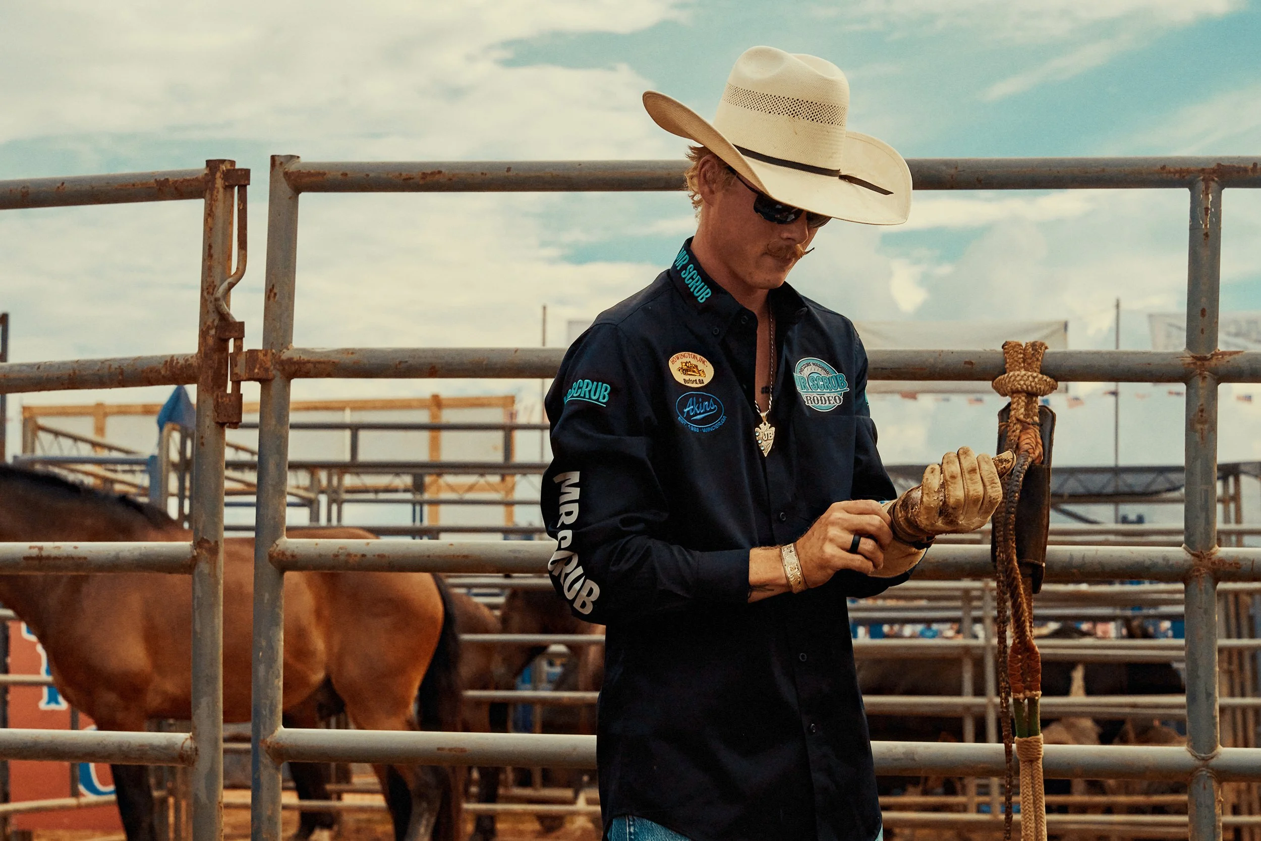 A man wearing a cowboy hat, sunglasses, and a dark shirt with rodeo patches checks his phone at a rodeo arena with horses and fencing in the background.