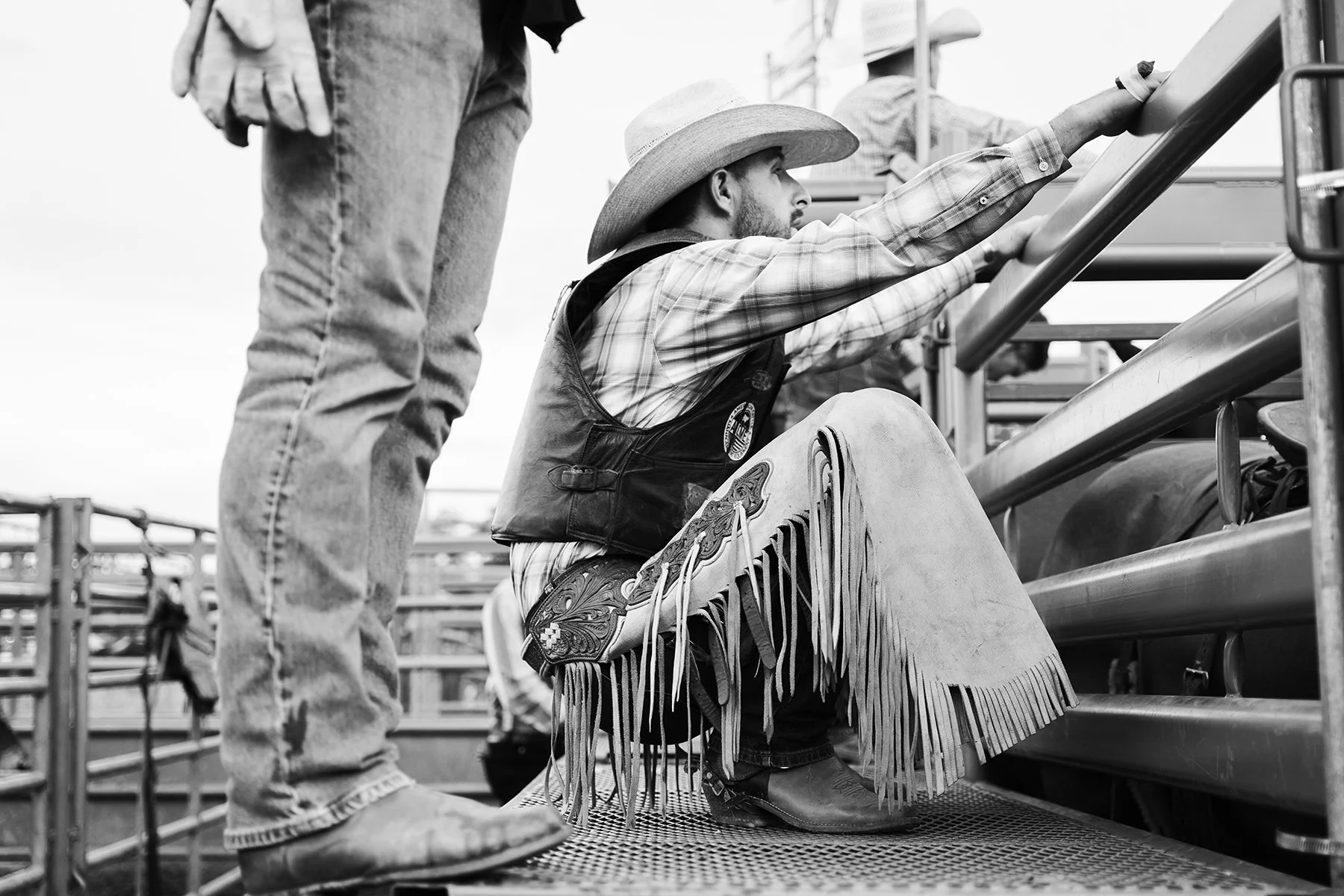 A man in cowboy attire, including a large sombrero, plaid shirt, and fringe chaps, reaching out to grab the railing. Another person standing nearby is shown from the waist down, dressed in jeans and boots.