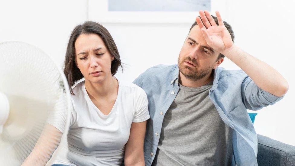 A couple sits in front of a fan, clearly struggling with the temperature