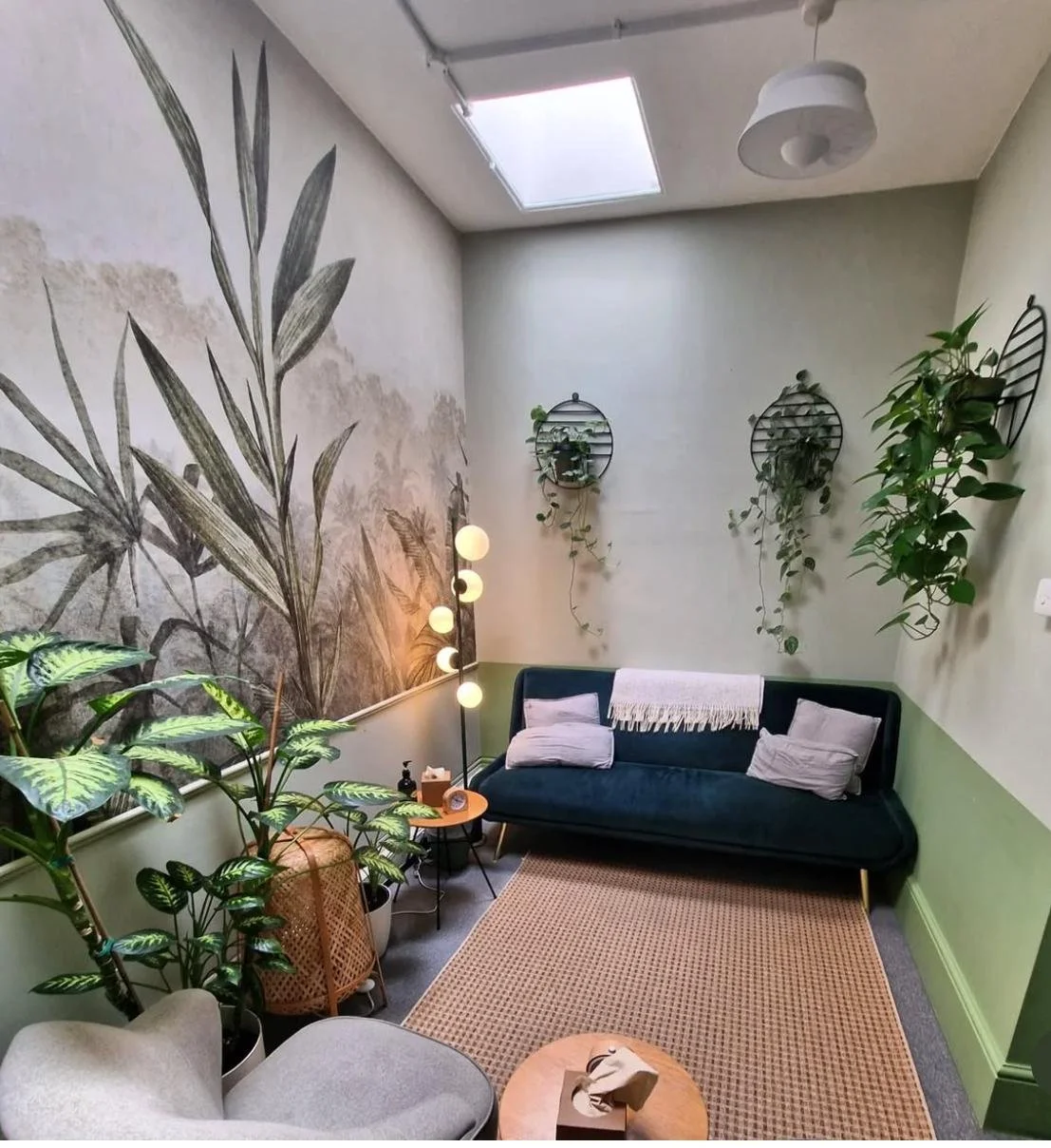 A cozy living room with a dark blue velvet couch, a small round wooden coffee table, a gray armchair, plants, wall art, and a skylight.