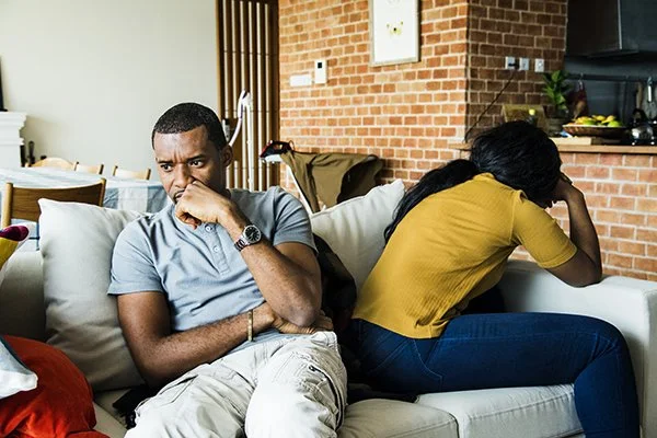 A couple sits back to back on a couch, both expressing sadness