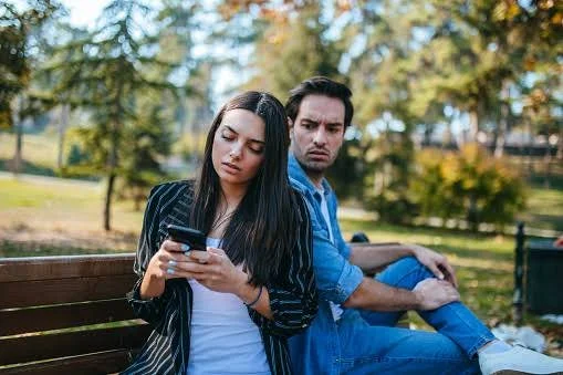 A woman is looking at her phone while her partner looks over her should suspiciously