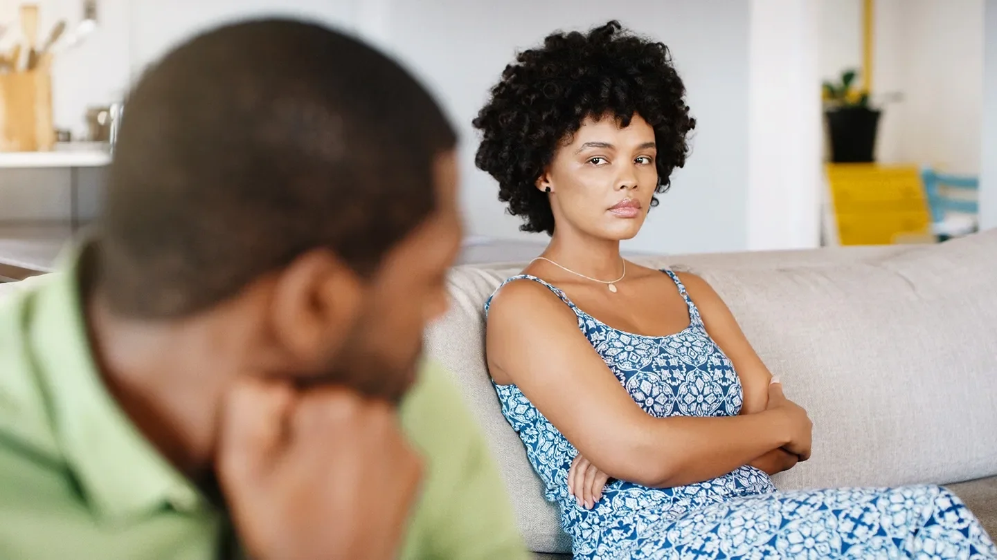 A man and a woman sit on a couch, she is in focus and he is blurred in the foreground. She is looking at him with sadness and pain.