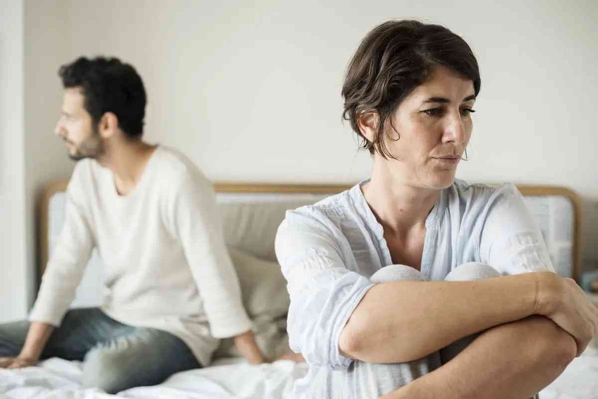A couple sit's on a bed facing different directions both seeming sad and deep in thought