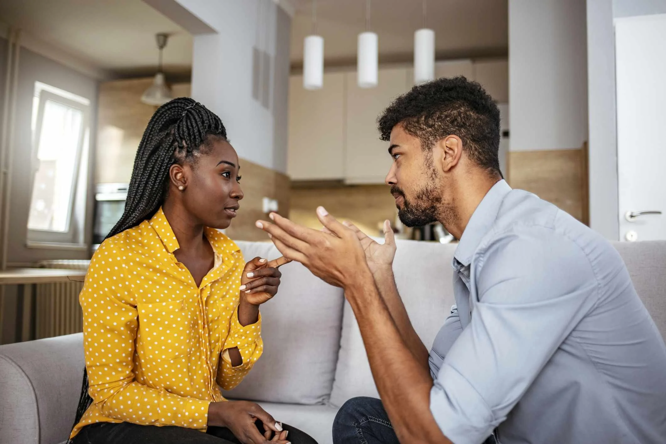 A couple sit on a couch engaged in conflict, with one partner pointing while the other holds their hands up
