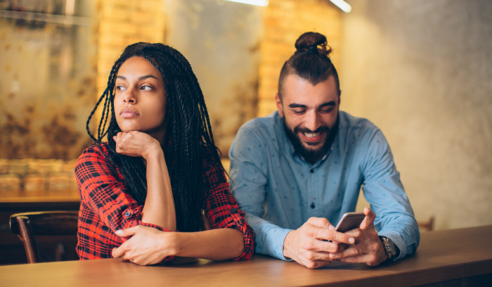 A woman is distant while a man sits beside her happily engaged with his mobile phone
