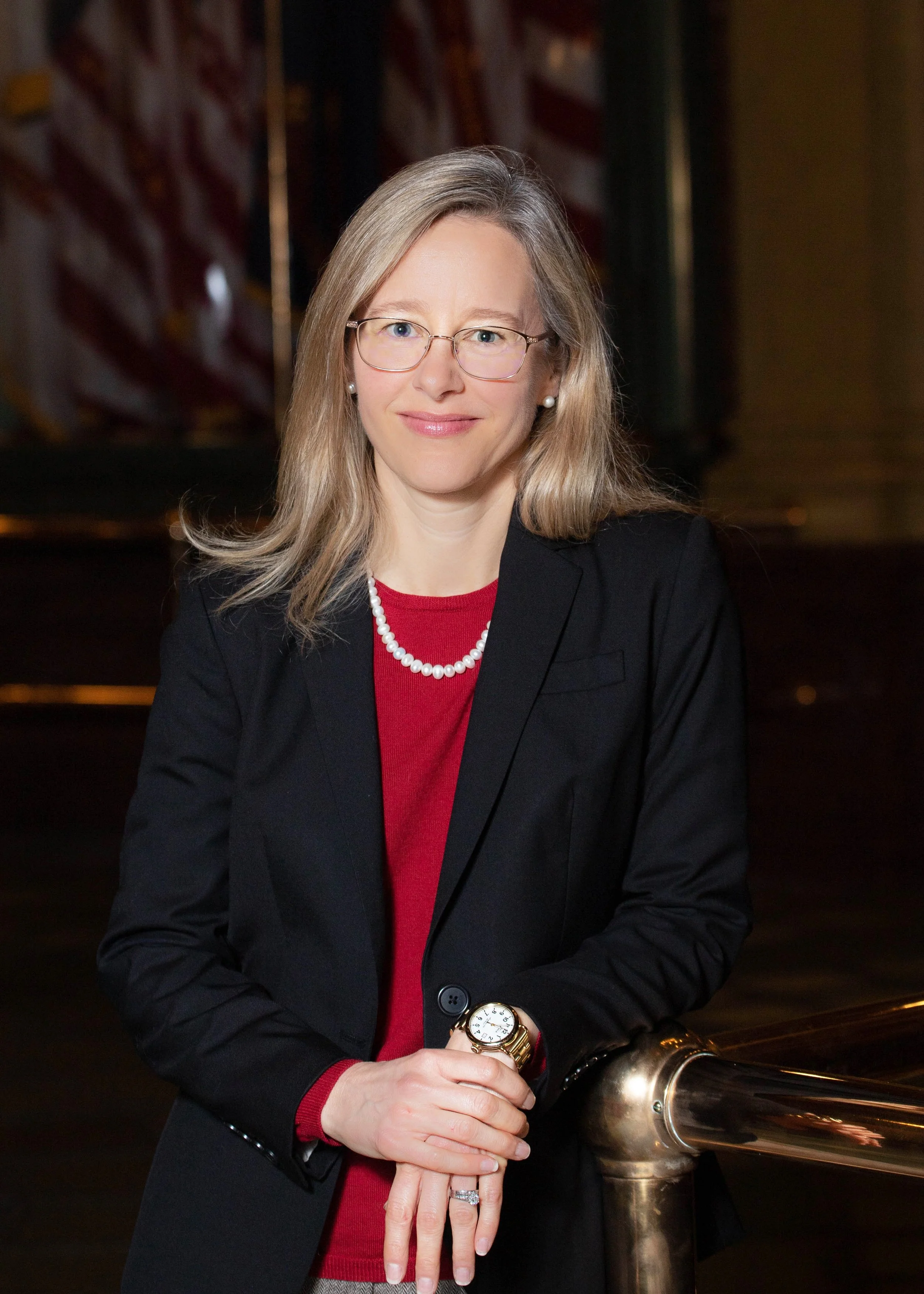 A professional woman with blonde hair, glasses, and pearl earrings, wearing a black blazer, red shirt, and pearl necklace, standing indoors with an American flag in the background.