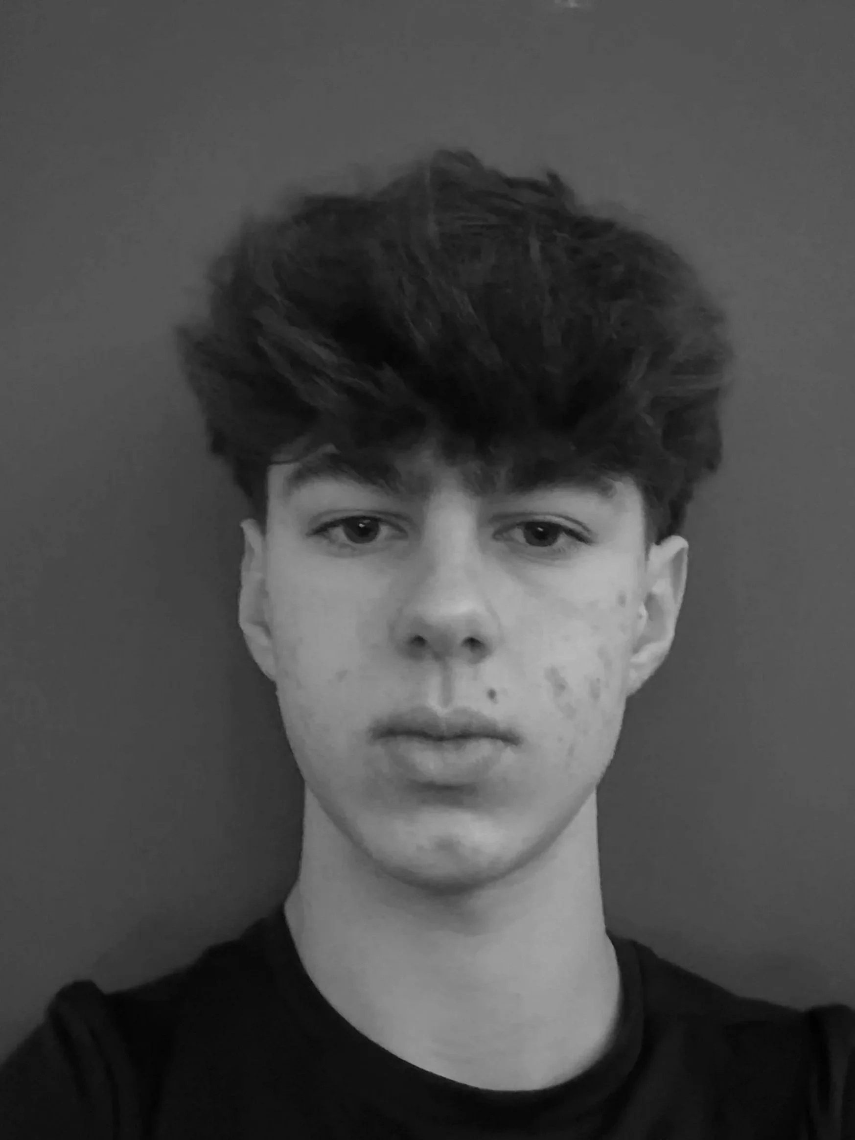Black and white portrait of a young male with tousled dark hair and fair skin, wearing a dark shirt, against a plain background.