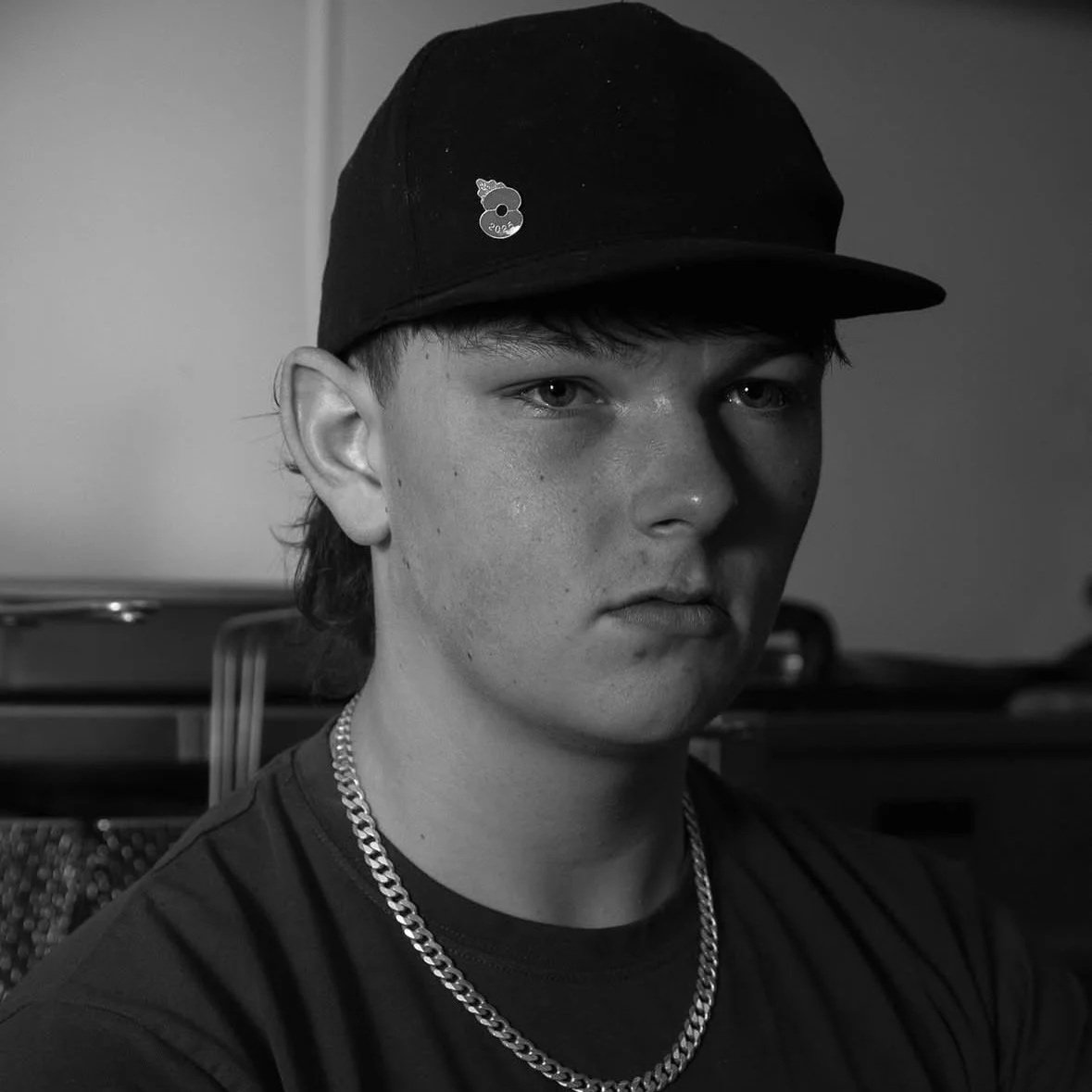 A young man wearing a black baseball cap with a small logo, a chain necklace, and a dark shirt. He has light skin, short hair, and is looking directly at the camera. The background appears to be indoors with some equipment behind him.