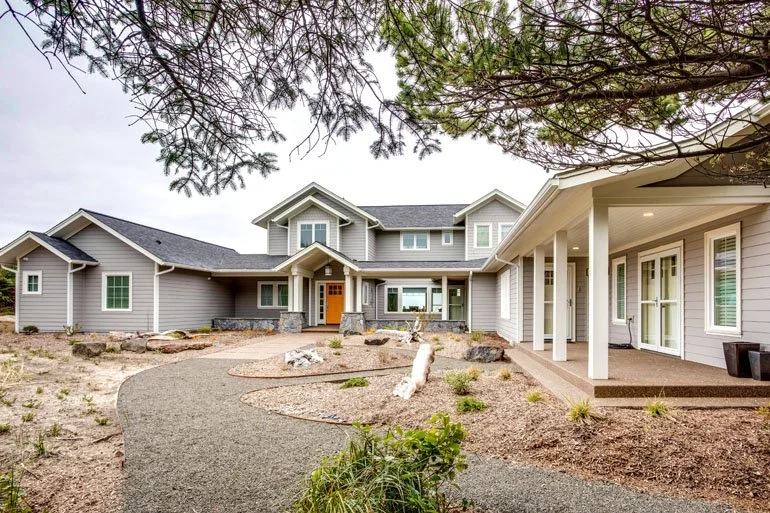 Front view of a large modern house with a gray exterior, multiple gables, and a covered patio area, surrounded by a sandy, landscaped yard with plants and trees.