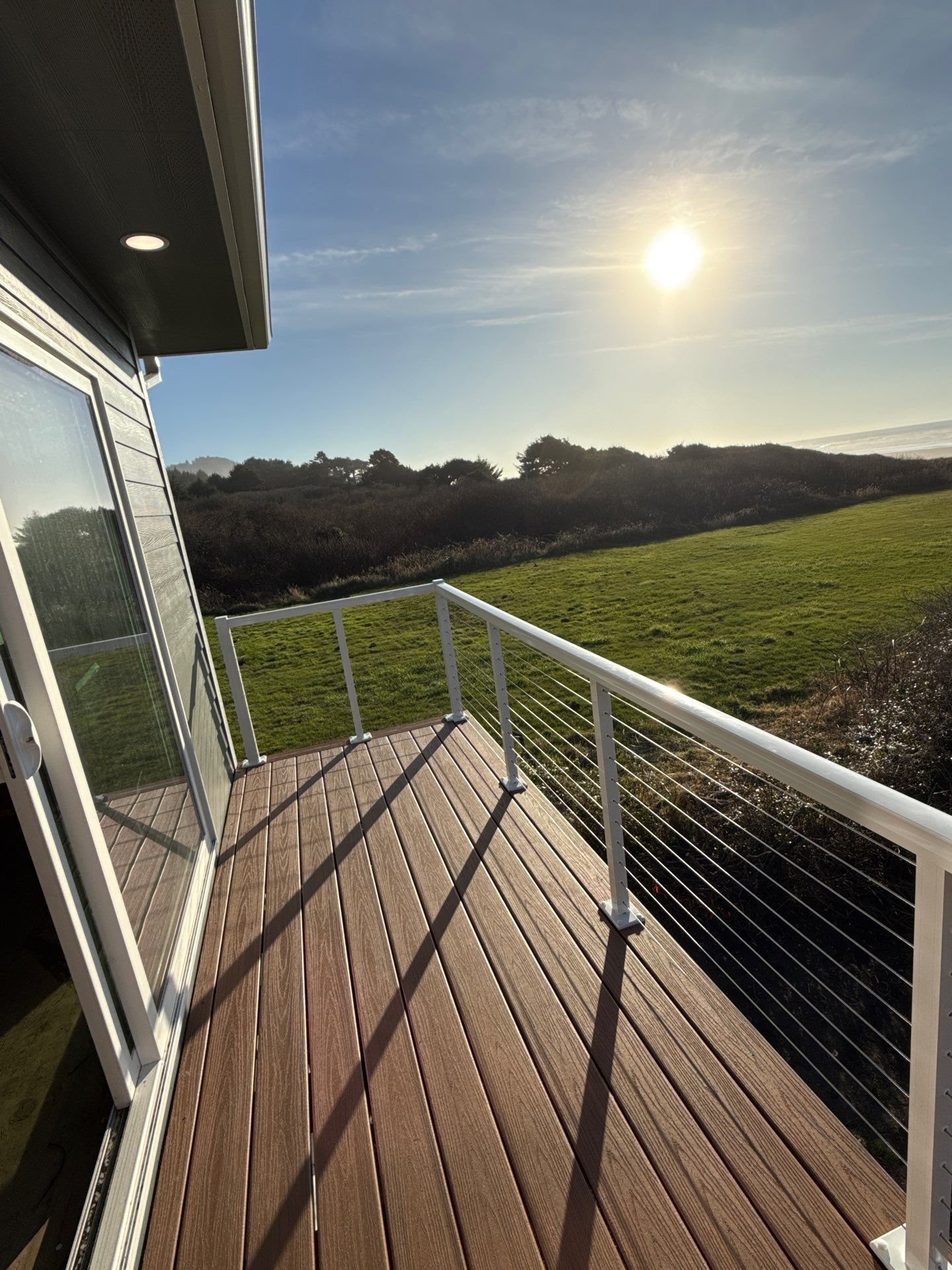 A scenic view from a balcony showing a grassy field, bushes, trees in the distance, with the sun shining in a mostly clear sky.