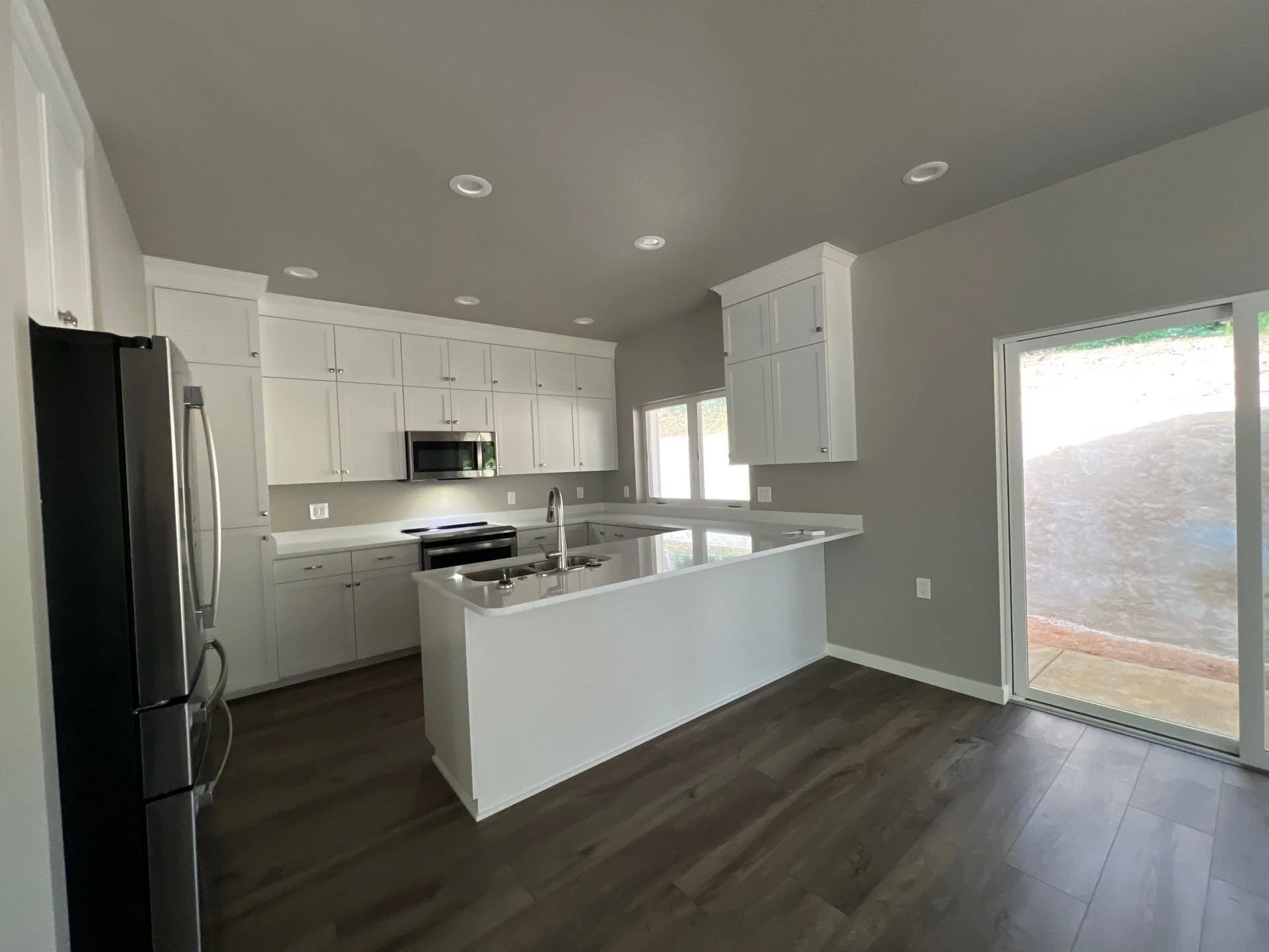 Modern kitchen with white cabinets, stainless steel refrigerator, microwave, stovetop, and a kitchen island with a sink, and a sliding glass door leading outside.