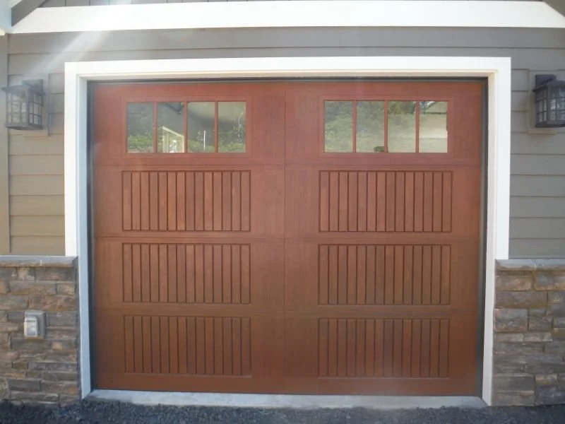 Wooden garage door with four rectangular windows at the top, framed by a white border, attached to a house with a stone and siding exterior.