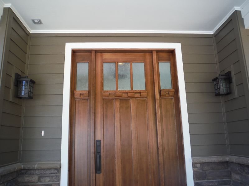 Front door made of wood with four upper glass panes, surrounded by gray siding and stone on the bottom, with two black outdoor wall lanterns on each side.