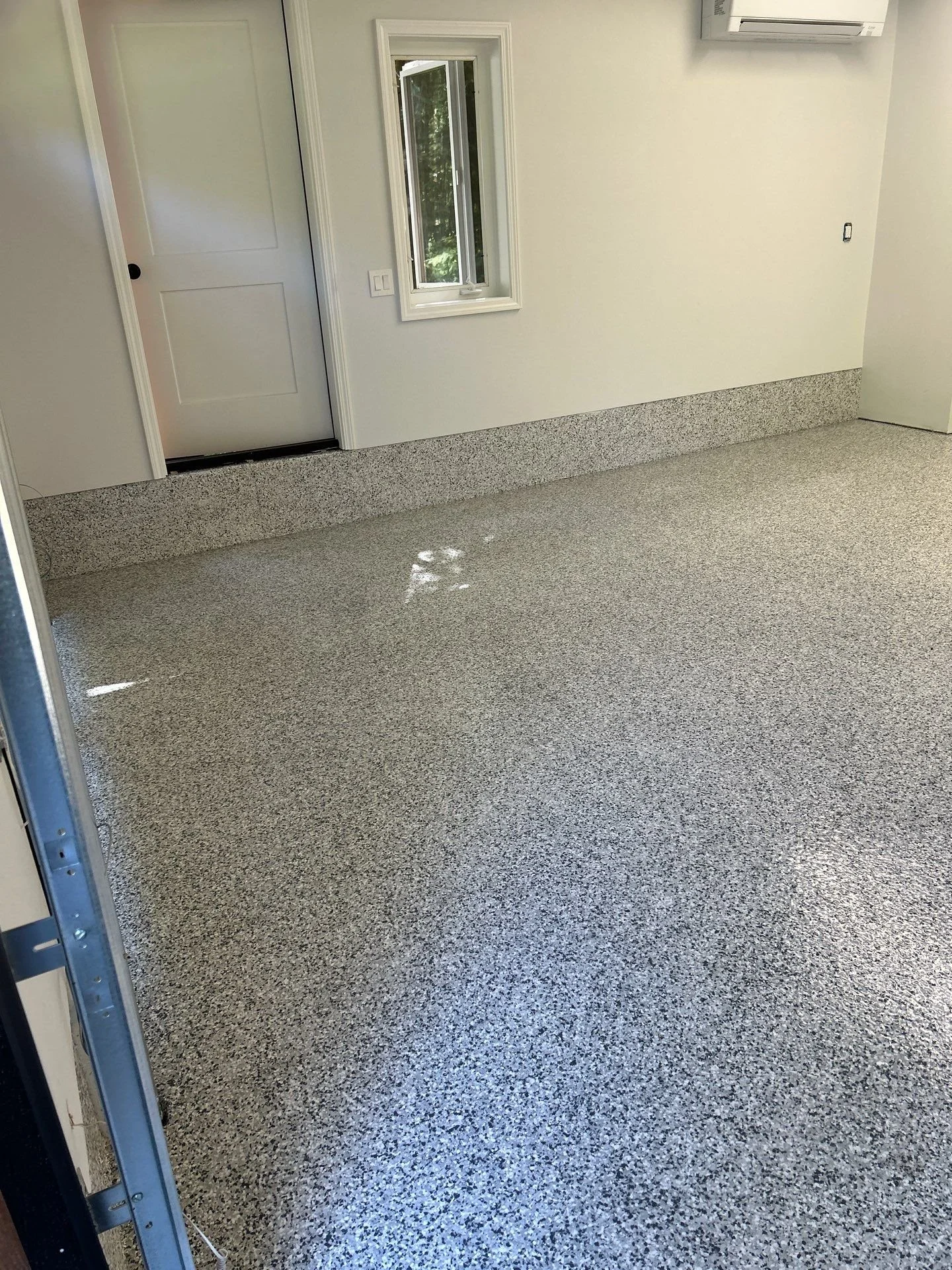 Empty garage with speckled gray epoxy flooring, a white door, a small window, and an air conditioning unit on the wall.
