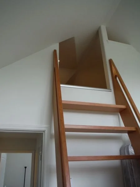 View of a loft area accessed by a wooden ladder inside a house with white walls.