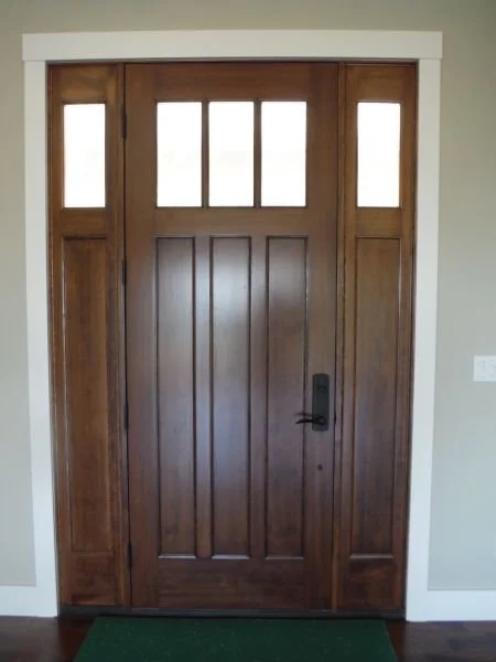 Wooden front door with four small frosted windows at the top, a black handle, and a lock, surrounded by white trim and a light-colored wall.