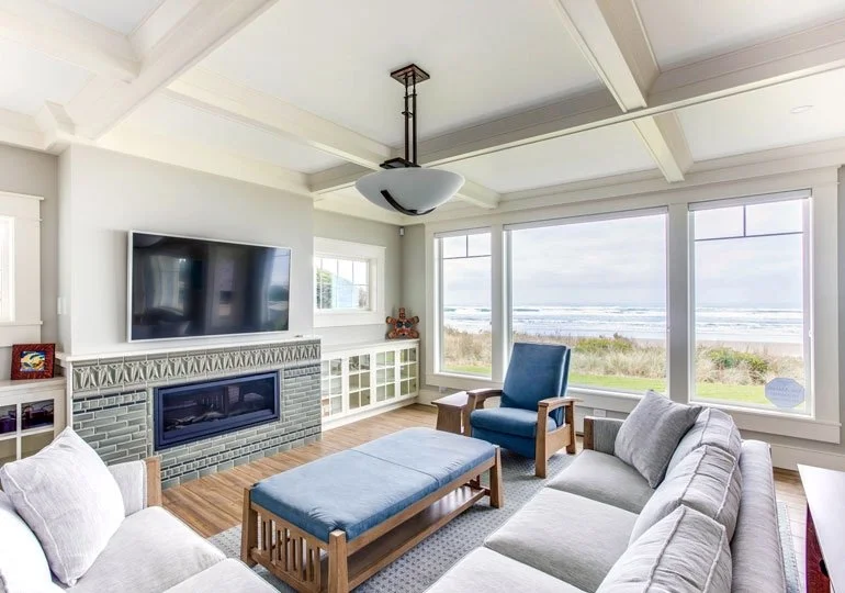 Living room with large windows overlooking the beach, a gray sectional sofa, a blue armchair, a fireplace with a TV above it, and a wood coffee table.
