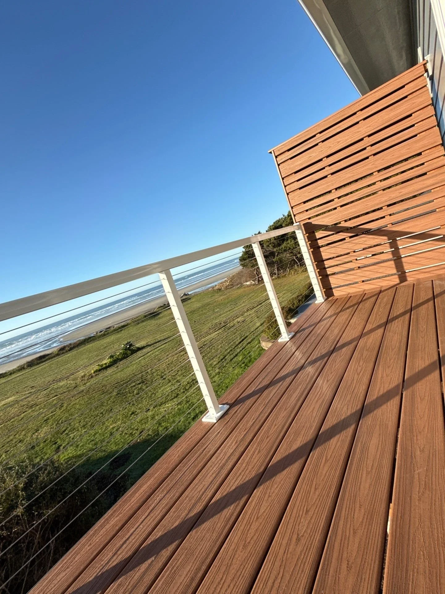 View of a wooden deck with a white railing overlooking a grassy area, trees, and the ocean under a clear blue sky.
