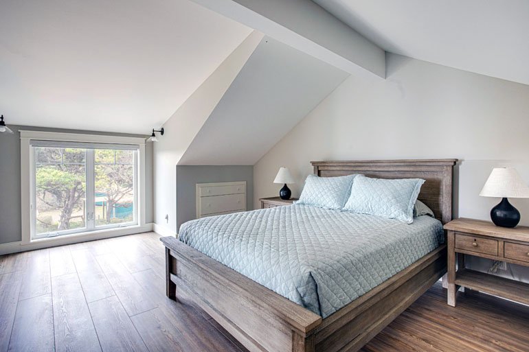Bedroom with wooden bed, light blue quilt, two matching pillows, two nightstands with black lamps, large window with a view of trees, sloped ceiling