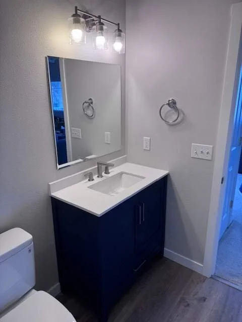 A modern bathroom with a navy blue vanity, white countertop, and rectangular mirror. Above the mirror, there is a light fixture with multiple bulbs. The wall is painted light gray, and there is a towel ring on the wall. A toilet is partially visible 
