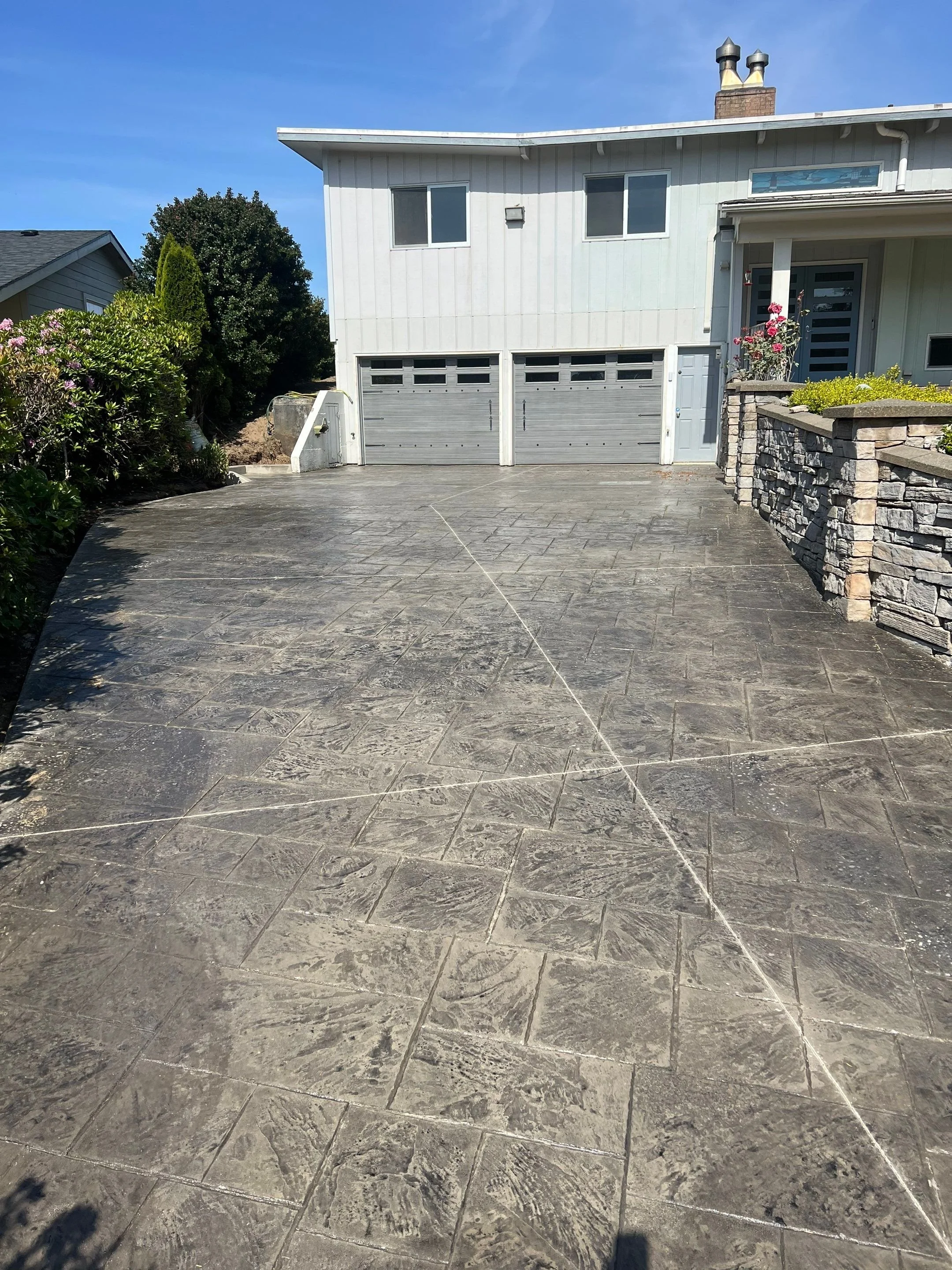 A concrete driveway in front of a modern house with a double garage door, a stone wall, and landscaping with plants and flowers, under a blue sky.