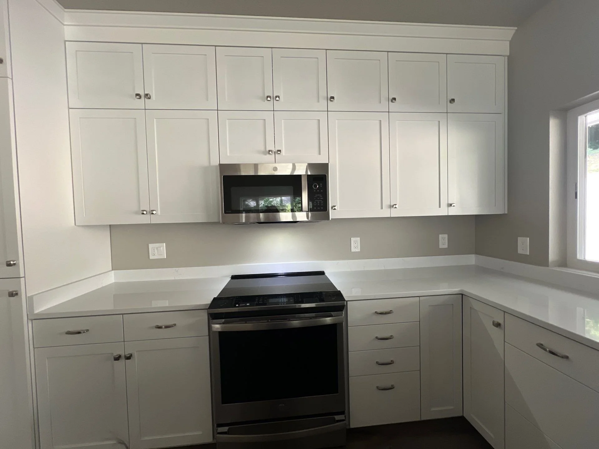 White kitchen cabinets with a microwave oven, stove, and white countertops under a window.