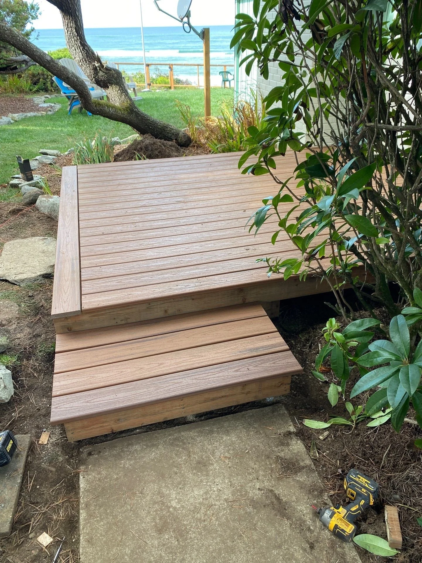 Newly constructed wooden deck with two levels in a backyard, surrounded by plants and overlooking the ocean, with tools on the ground nearby.