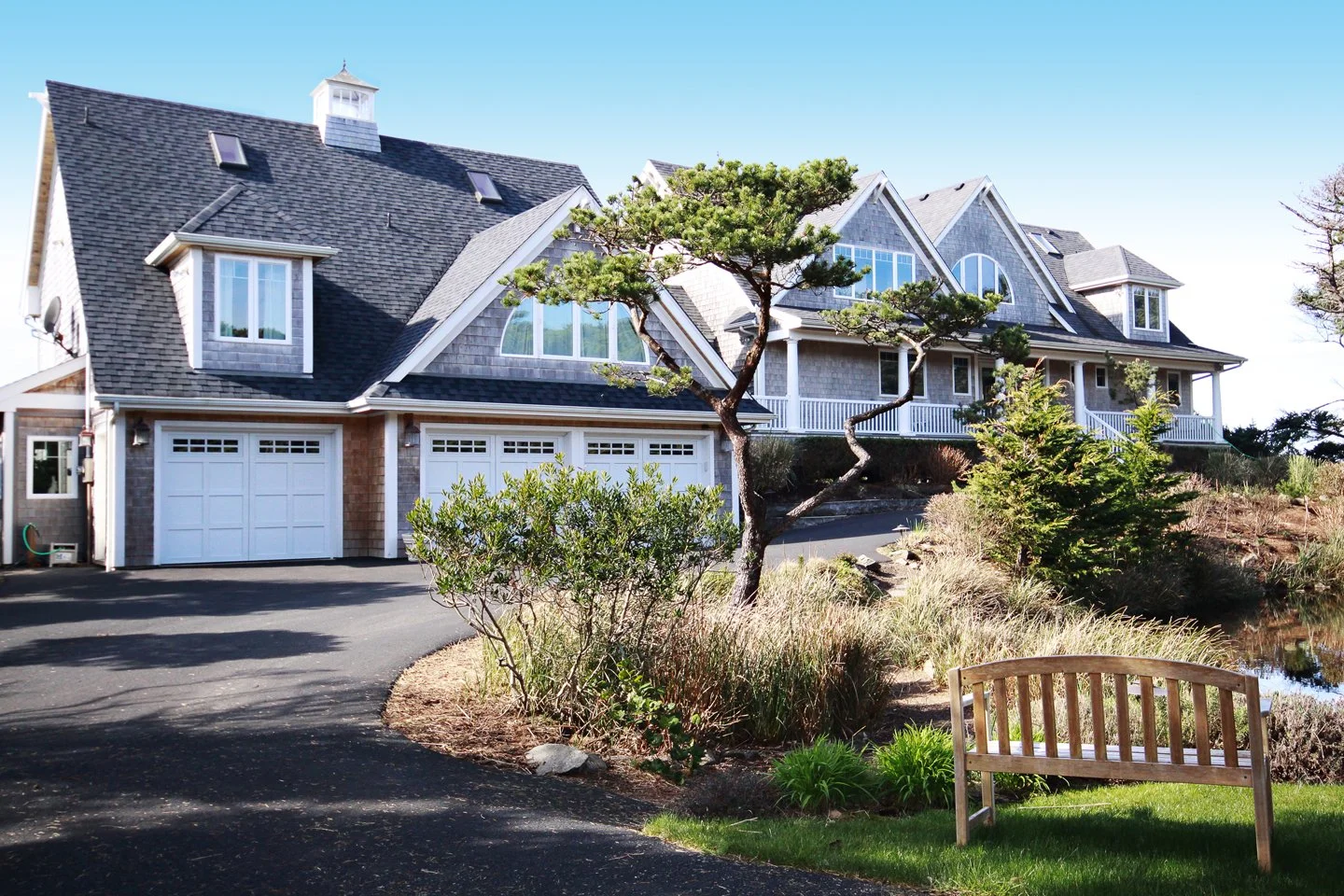 Large house with gray siding, multiple gabled roofs, and a three-car garage. Front yard with trees, shrubs, a pond, and a wooden bench.