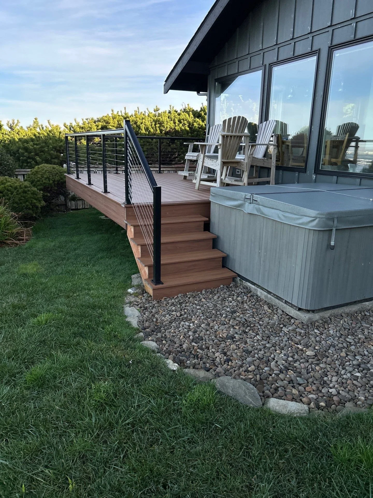 A raised wooden deck with black railing attached to a house with large glass windows. The deck has outdoor chairs and leads down to a grassy yard with trees. A hot tub is positioned next to the house beneath the deck.