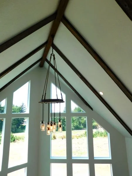 Interior of a house with an angled ceiling, wooden beams, and large windows showing greenery outside. A modern chandelier with multiple light bulbs hangs from the ceiling.