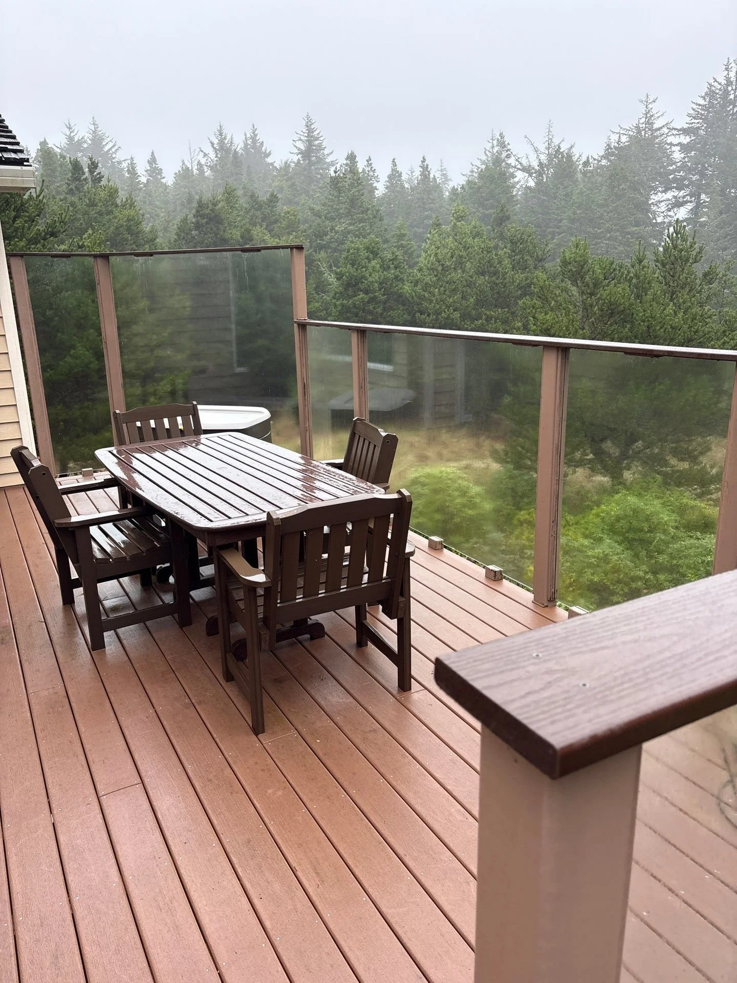 Wooden outdoor balcony with a wet table and four matching chairs, surrounded by a glass railing, overlooking a forested area on a rainy day.