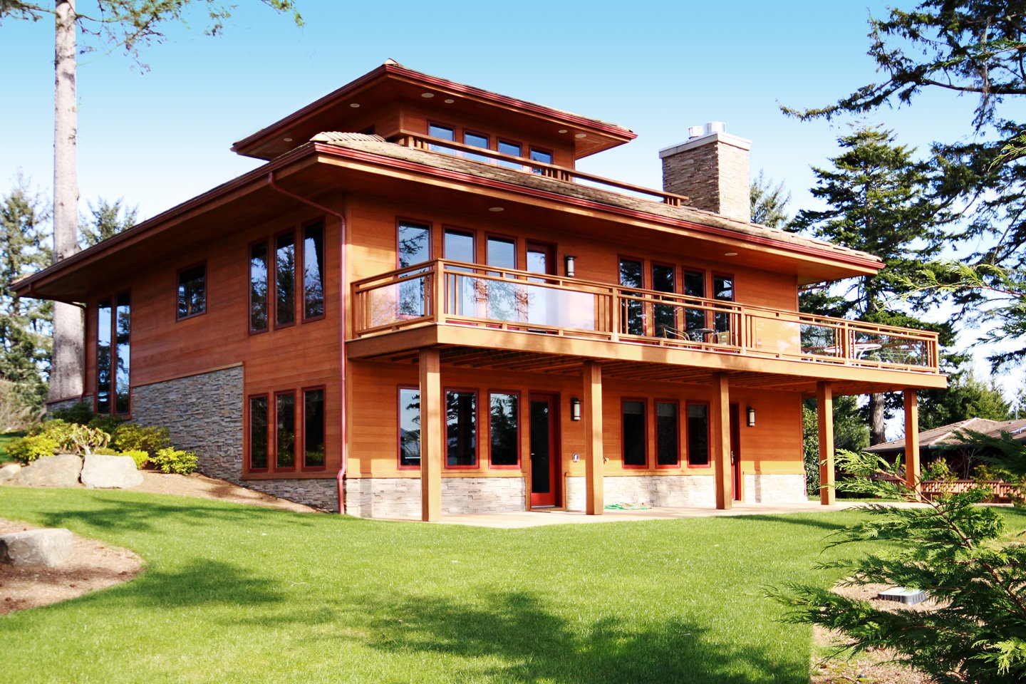 A large modern wooden house with multiple windows, balcony, and stone accents, surrounded by a well-maintained green lawn and trees.