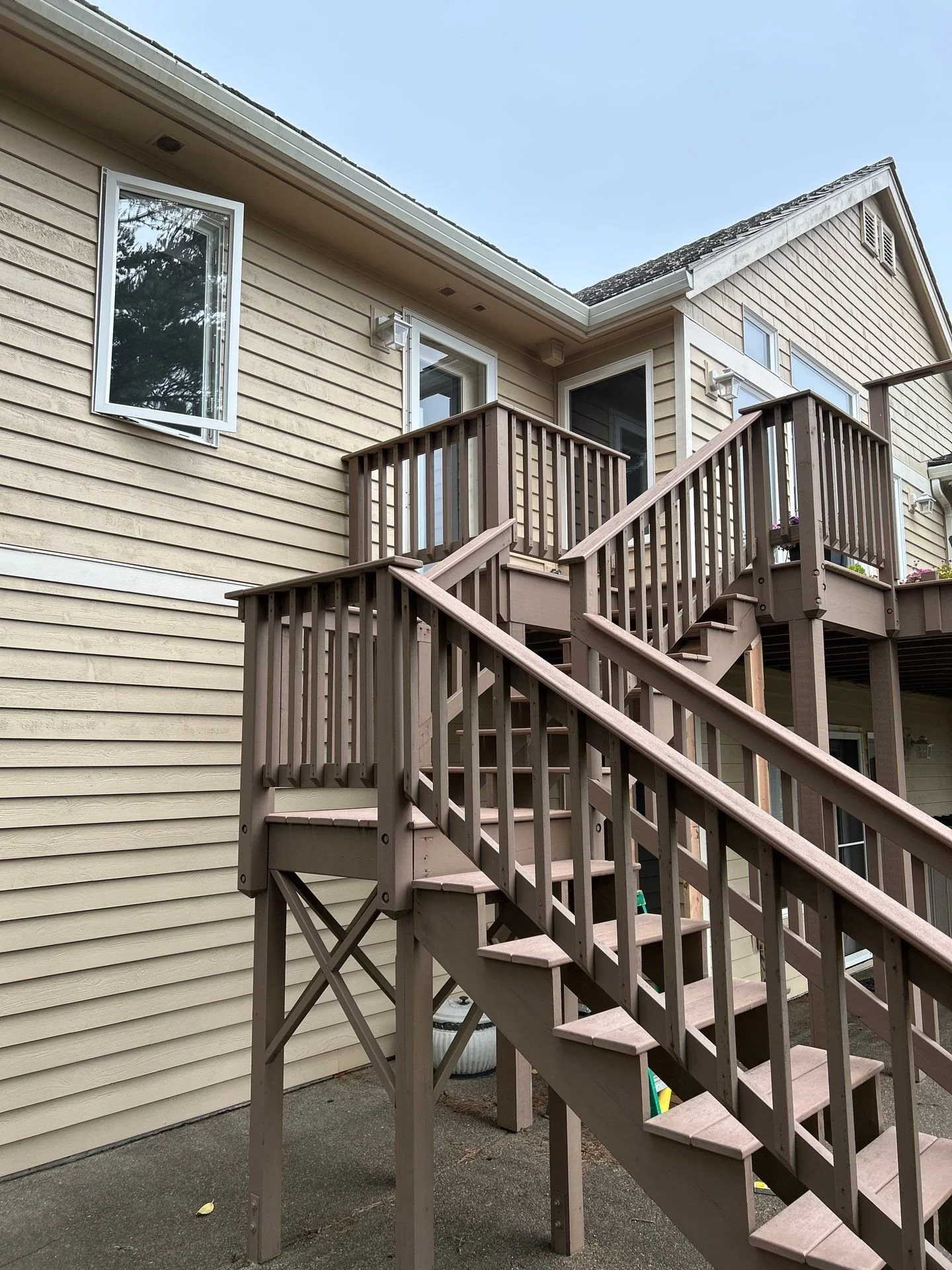 Back exterior view of a house with beige siding, featuring an outdoor staircase leading to a second-story deck with brown railings. The house has multiple windows, and the deck has a small flower pot on the railing.