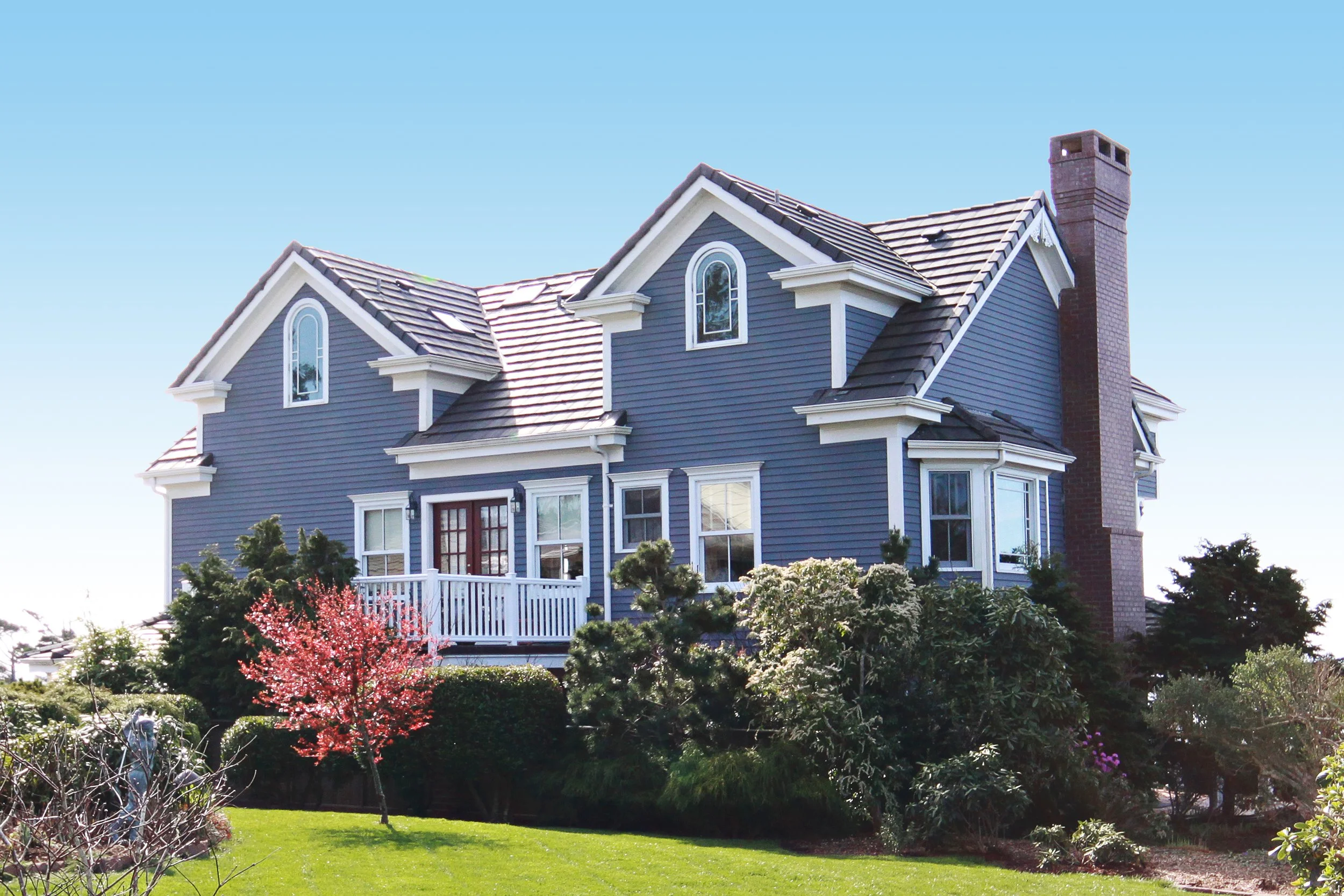 Blue two-story house with gabled roof, multiple windows, front porch with white railing, surrounded by trees and a well-kept lawn, under a clear blue sky.
