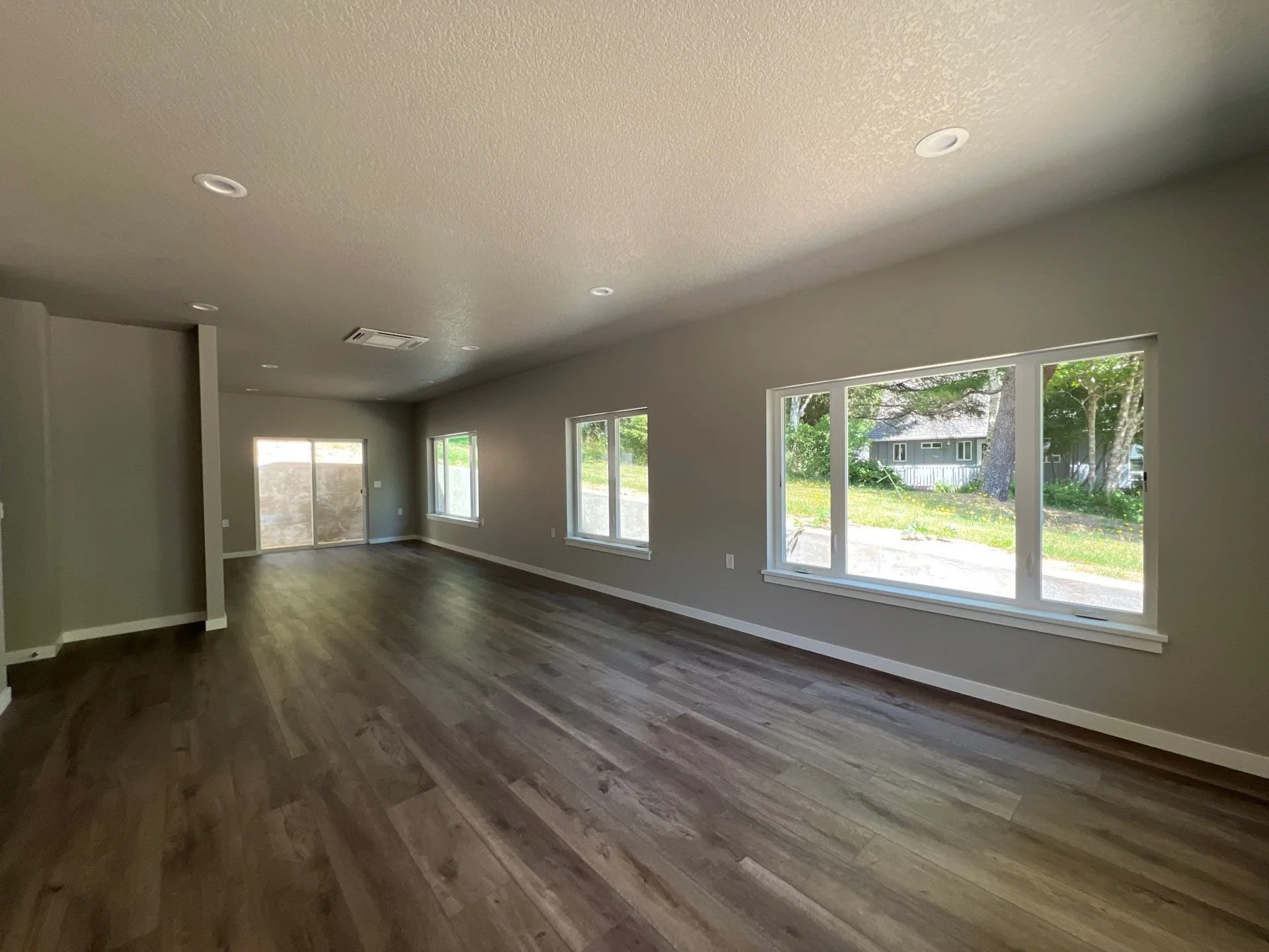 Empty living room with large windows, wood flooring, and neutral-colored walls, with sunlight streaming in.