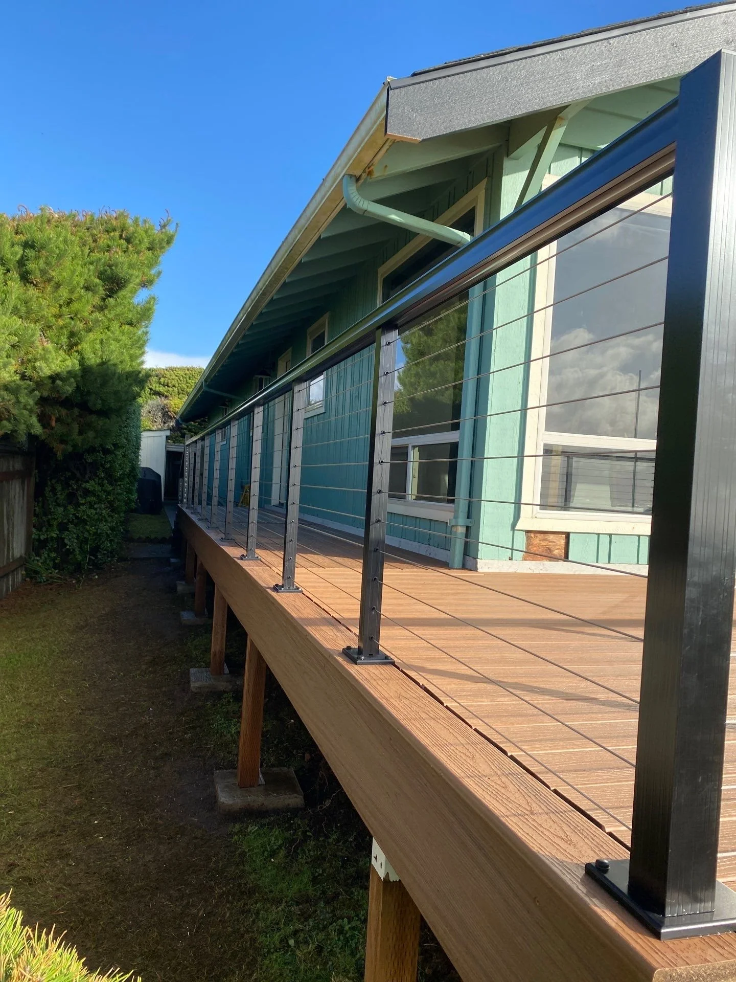 Wooden deck with metal railing in front of a blue house with green trim and white window frames, under a clear blue sky.