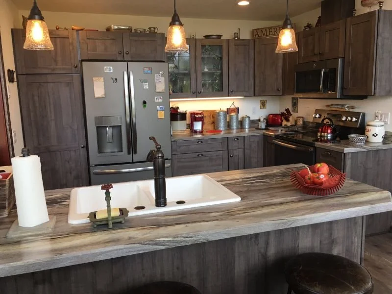 Modern kitchen with gray wooden cabinets, stainless steel refrigerator, microwave, and black stove; pendant lights hanging above a wooden kitchen island with a white sink and black faucet; red fruit bowl on the island.