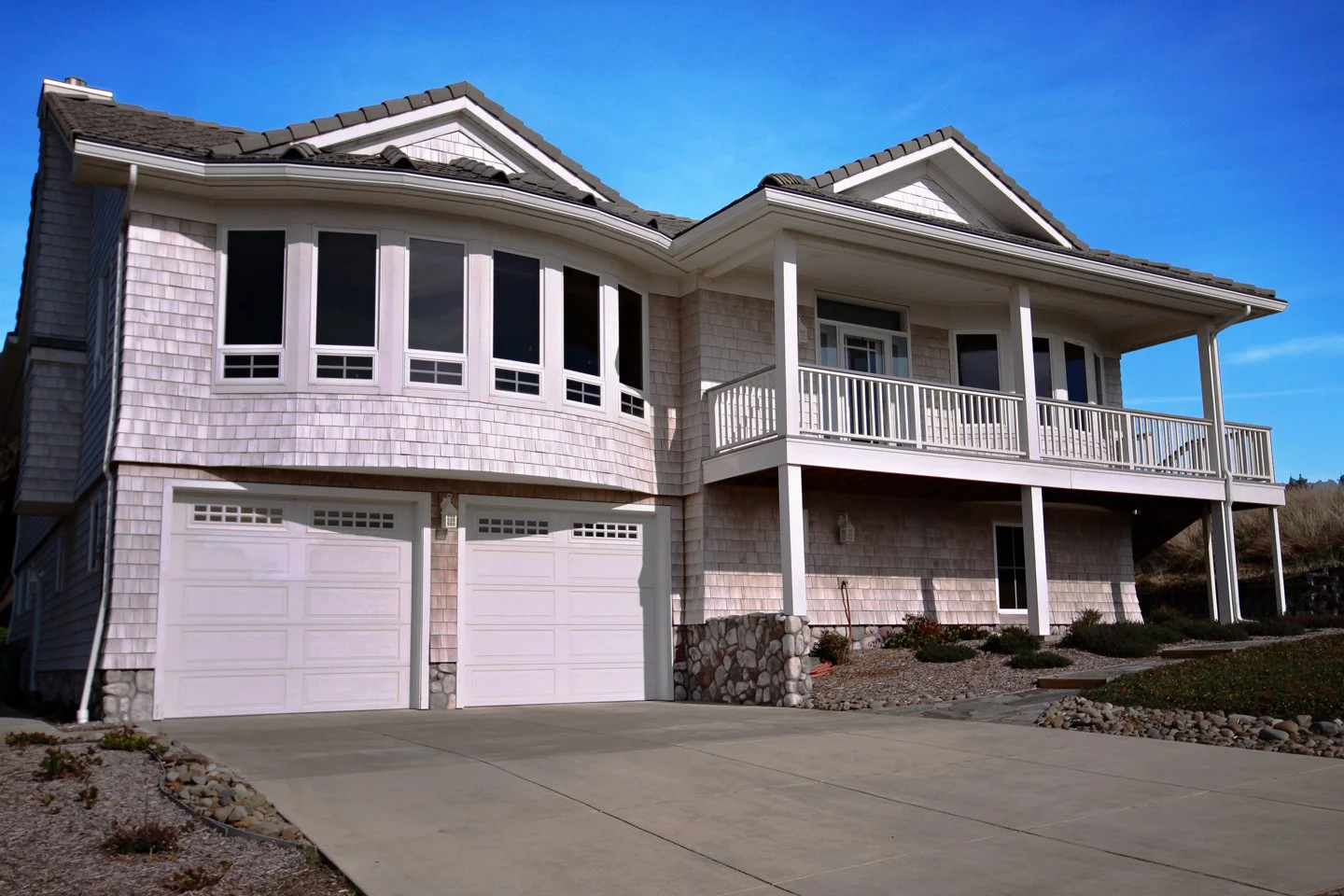 A large, modern two-story house with a white garage door, a spacious front porch with white railings, and multiple windows. The house has a sloped roof and beige exterior siding, with a driveway leading to the garage and a landscaped front yard.