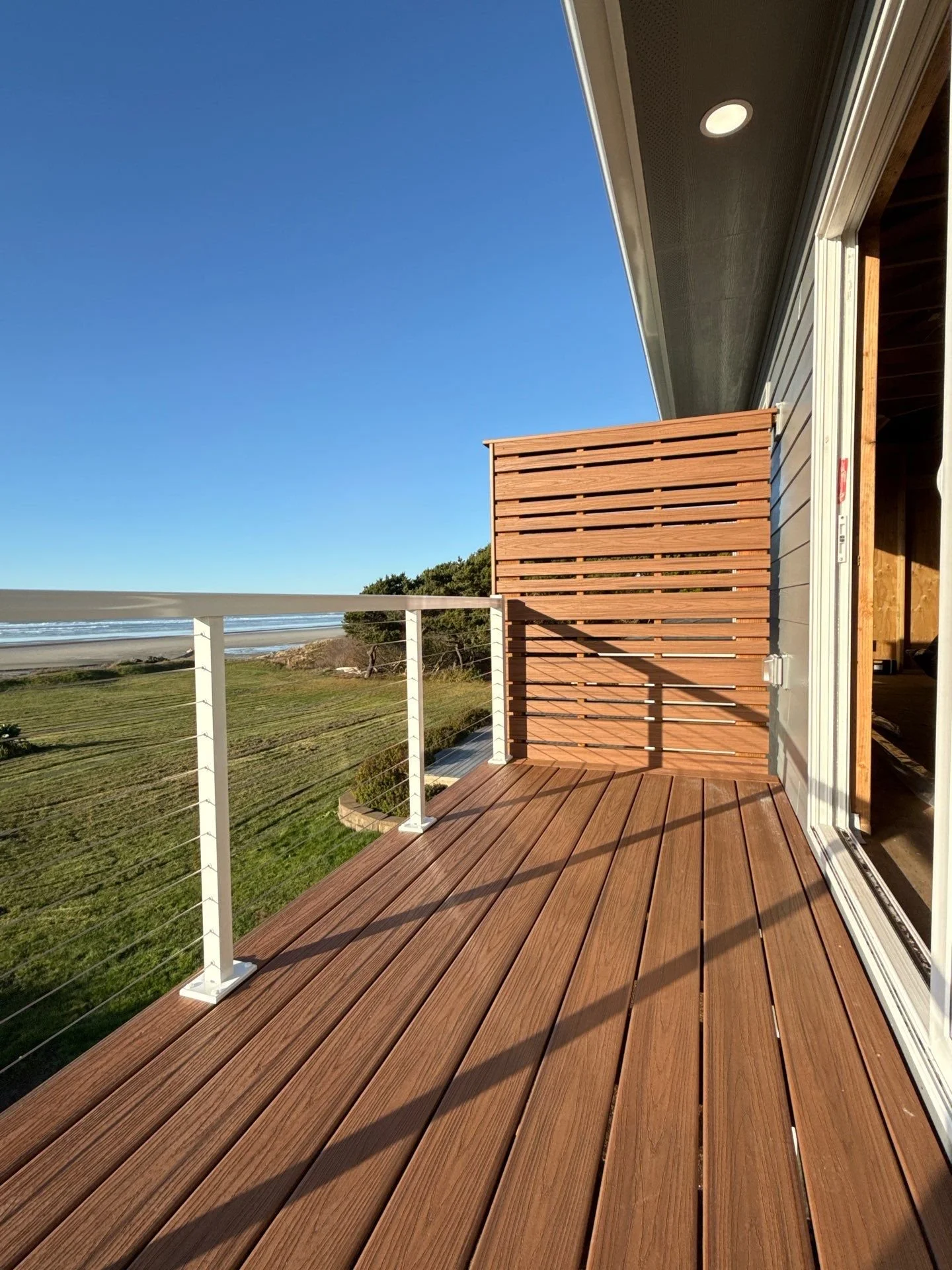 View of a wooden balcony with railing overlooking green fields and ocean, under a clear blue sky with sunlight casting shadows.